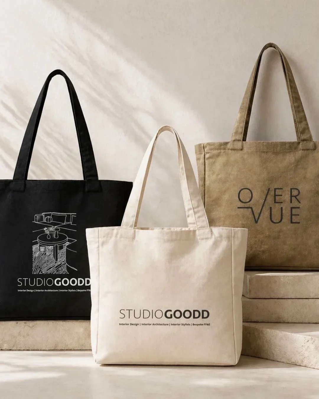 Three custom branded canvas tote bags in black, cream, and olive green, featuring printed logos for Studio Goodd and Overvue, displayed on stone steps for promotional merchandise inspiration.