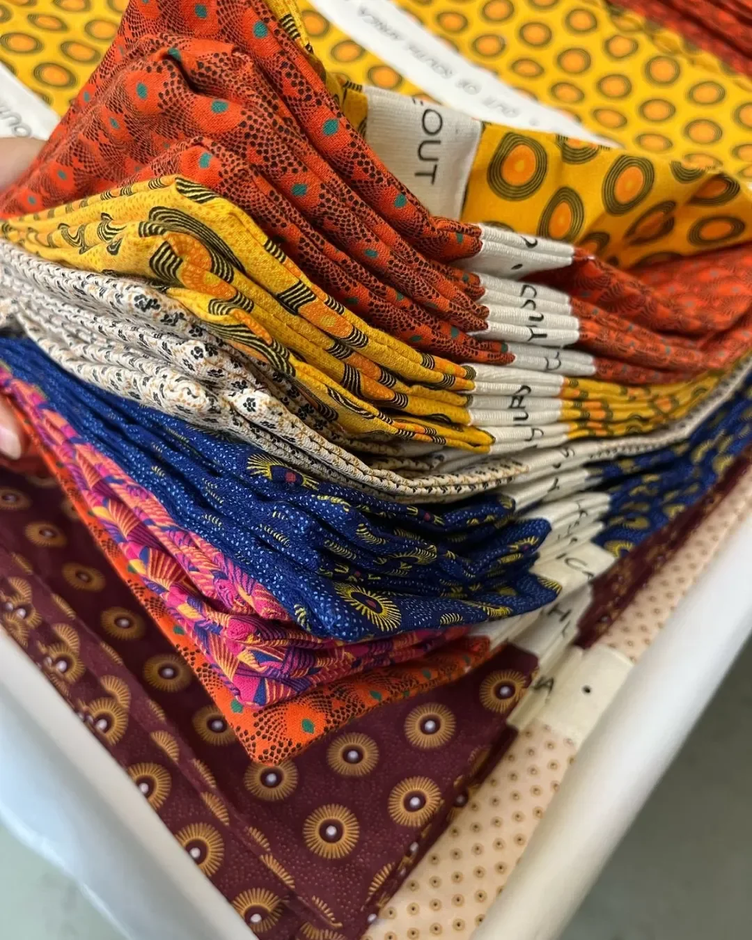 A high-angle close-up of a vibrant stack of folded 100% shweshwe tote bags in various patterns—including orange, yellow, blue, and maroon—each featuring a custom white cotton ribbon with 'OUT OF SOUTH AFRICA' branding, handmade in Cape Town by Soli.