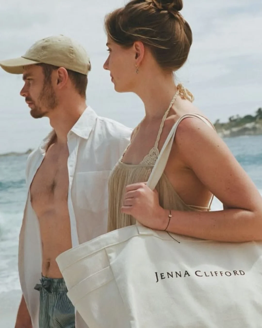 A woman walking on a beach carrying a large cream-colored 100% cotton canvas Weekending Bag with 'JENNA CLIFFORD' branding, Handmade by Soli in Cape Town for merchandise or corporate gifts.