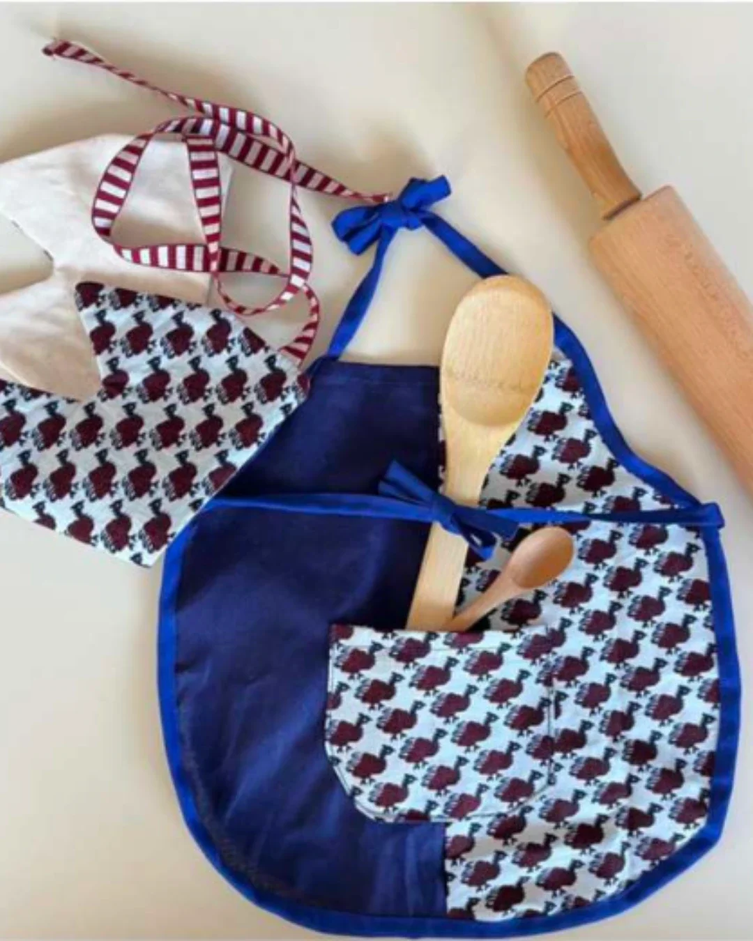 A handmade blue and white pattern Shweshwe kiddies apron with a dark blue pocket holding wooden kitchen utensils, crafted by Soli in Cape Town for sustainable hotel guest amenities.