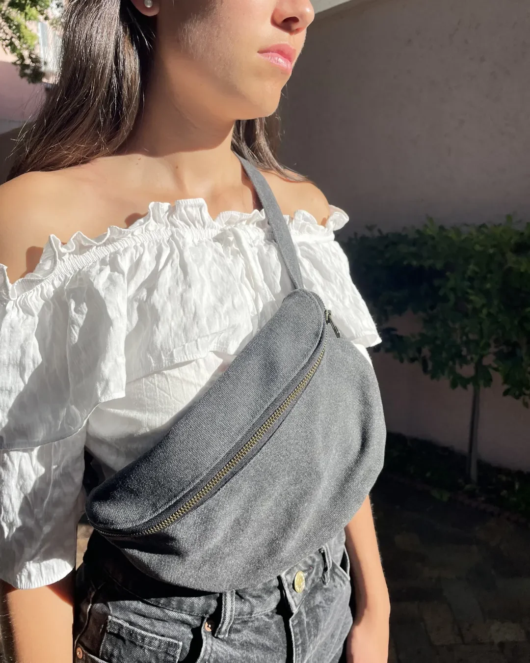 A person wearing a charcoal grey handmade cotton canvas moon bag across a white off-the-shoulder top, showcasing the practical and trendy fanny pack design crafted in Cape Town by Soli for corporate gifts and merchandise.
