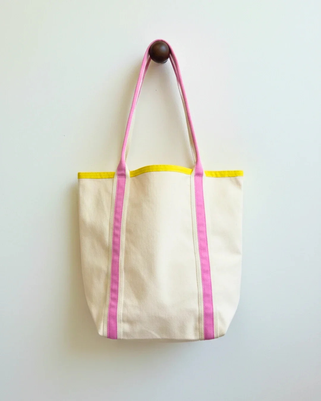 Cream heavy-weight cotton canvas Promenade bag hanging on a dark wooden peg, featuring bright pink vertical straps and a sunny yellow top trim, handmade by Soli in Cape Town for sustainable merchandise or corporate gifts.