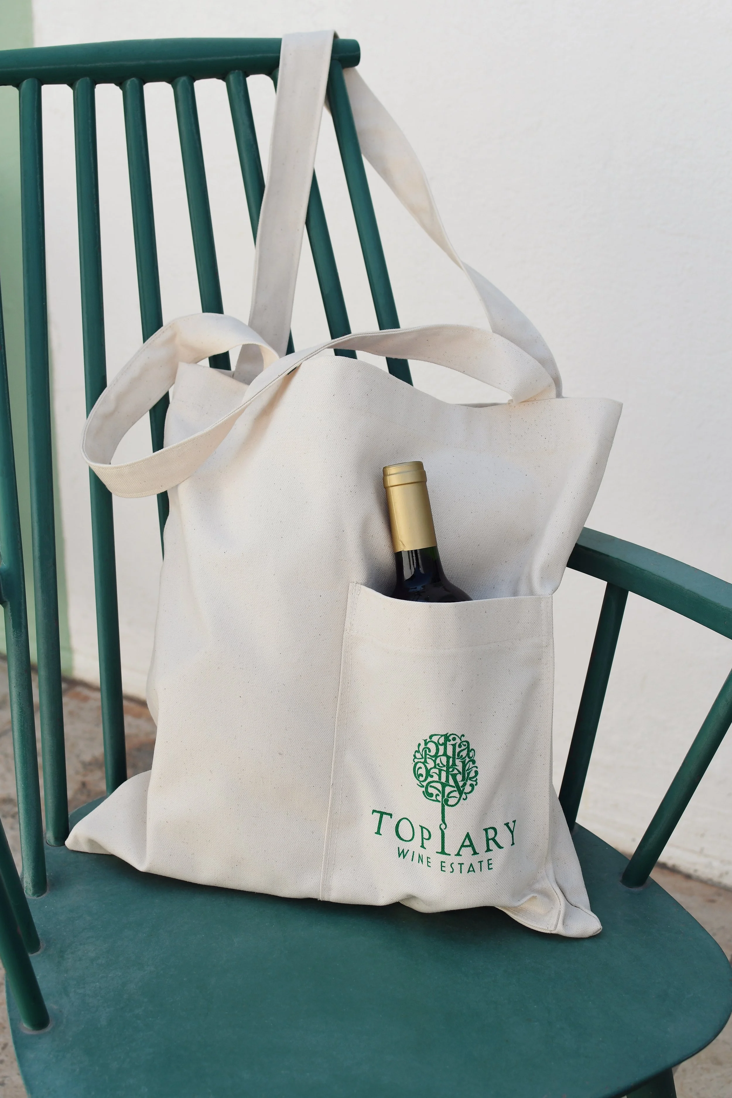 Canvas tote bag with "Topiary Wine Estate" logo on green chair, containing wine bottle.