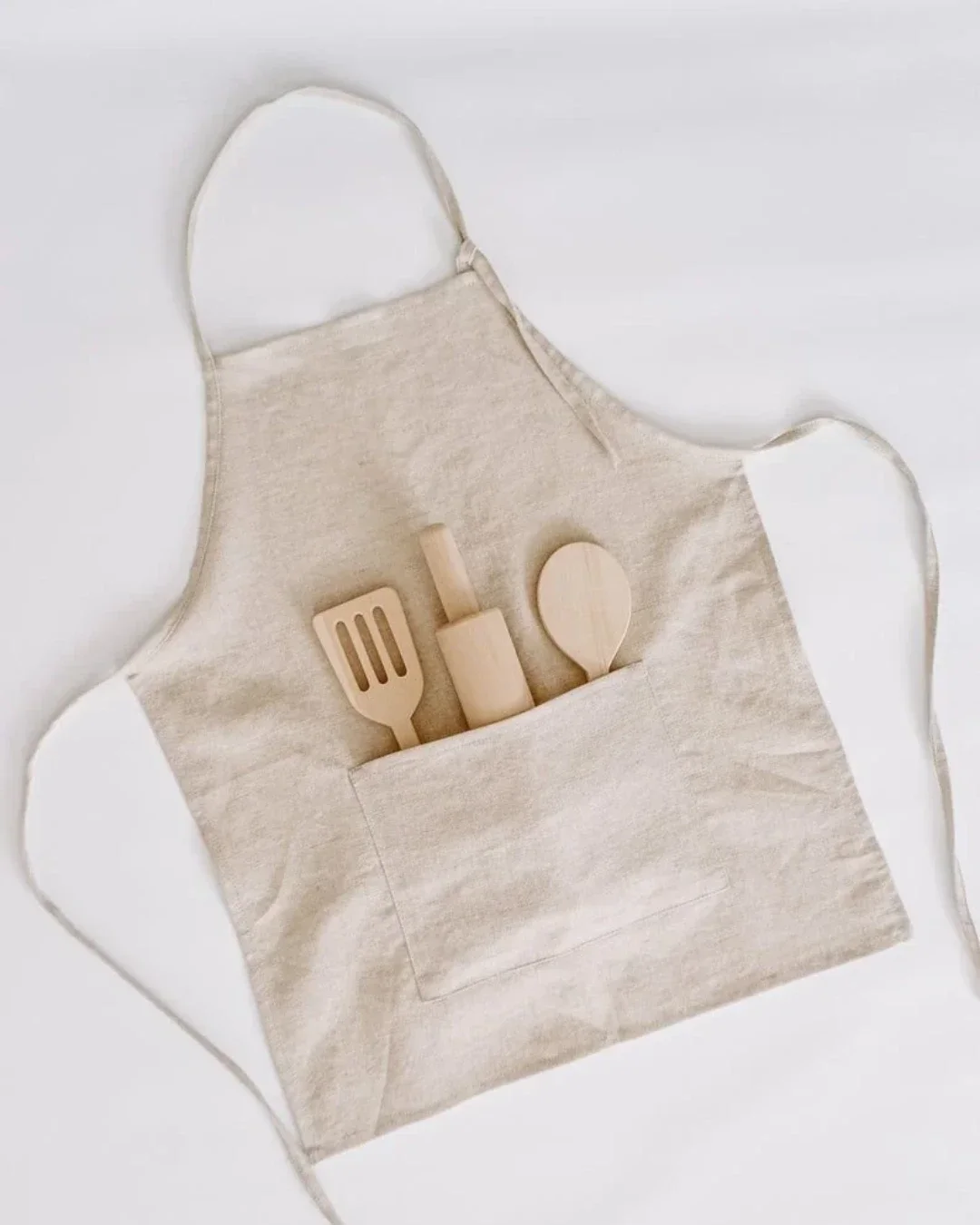 A beige handmade canvas kiddies apron with a large front pocket holding wooden toy kitchen utensils, designed by Soli in Cape Town for sustainable hotel amenities.