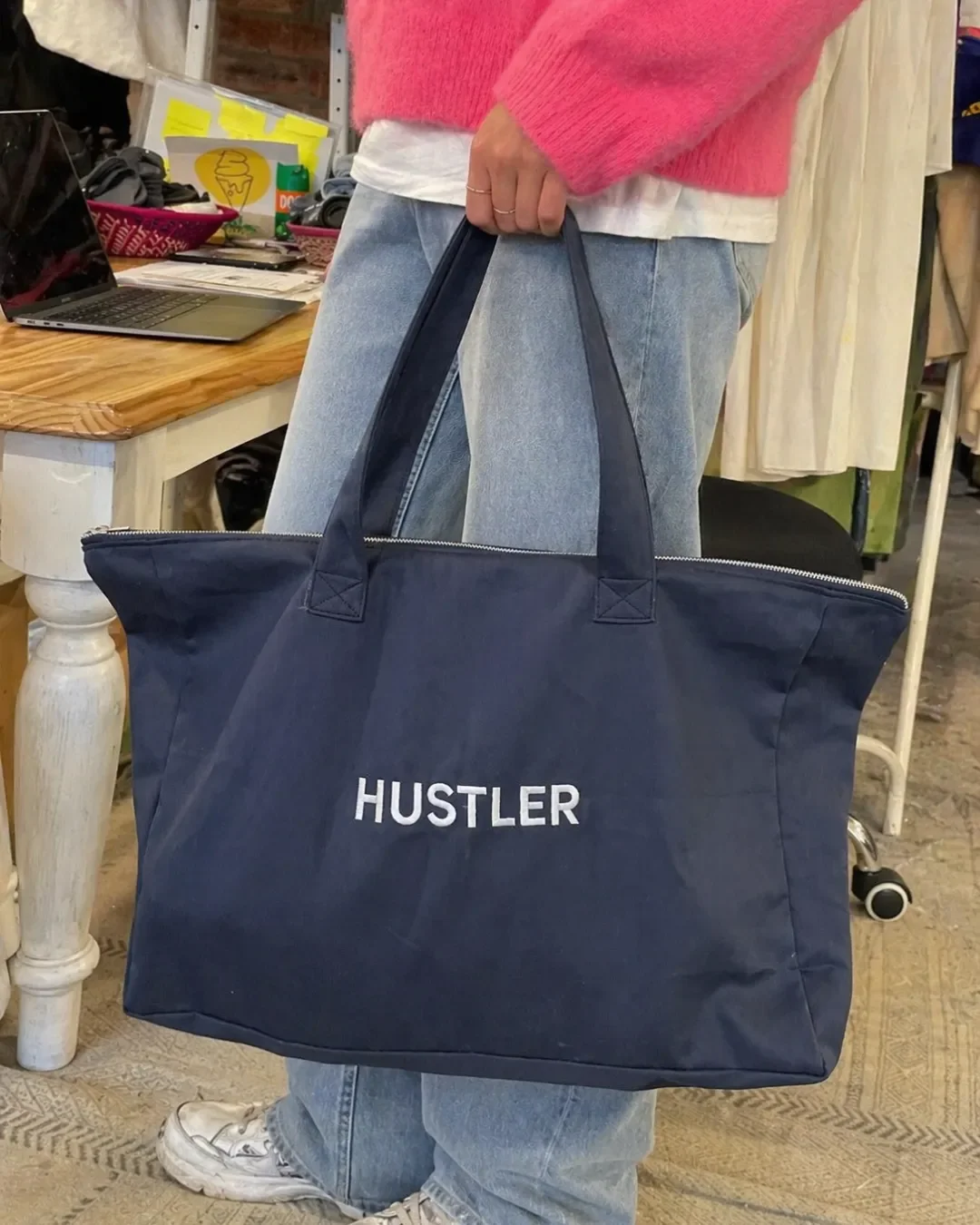 A large navy blue canvas tote bag branded with the word "HUSTLER" in bold white lettering, carried over the shoulder by a person in a bright pink sweater, within a creative studio environment for promotional merchandise.
