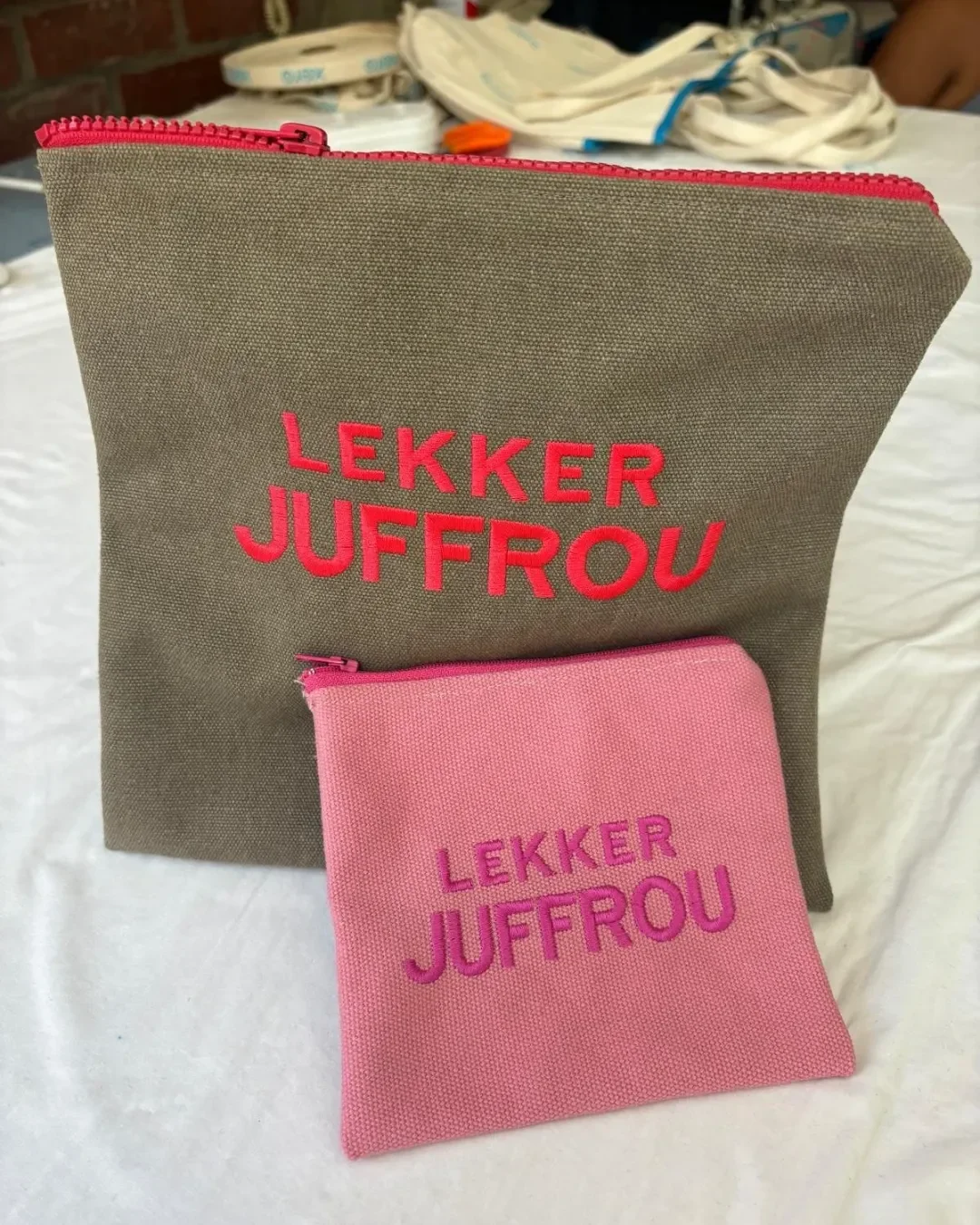 Two canvas pouches in olive green and pink with Lekker Juffrou embroidery and pink zippers on a white surface, Handmade by Soli in Cape Town for sustainable merchandise or corporate gifts.