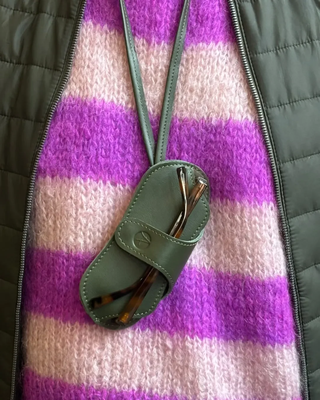 Close-up of an olive green leather sunglass holder pouch with a matching strap and debossed logo, holding tortoise shell glasses, custom made by Soli in Cape Town.