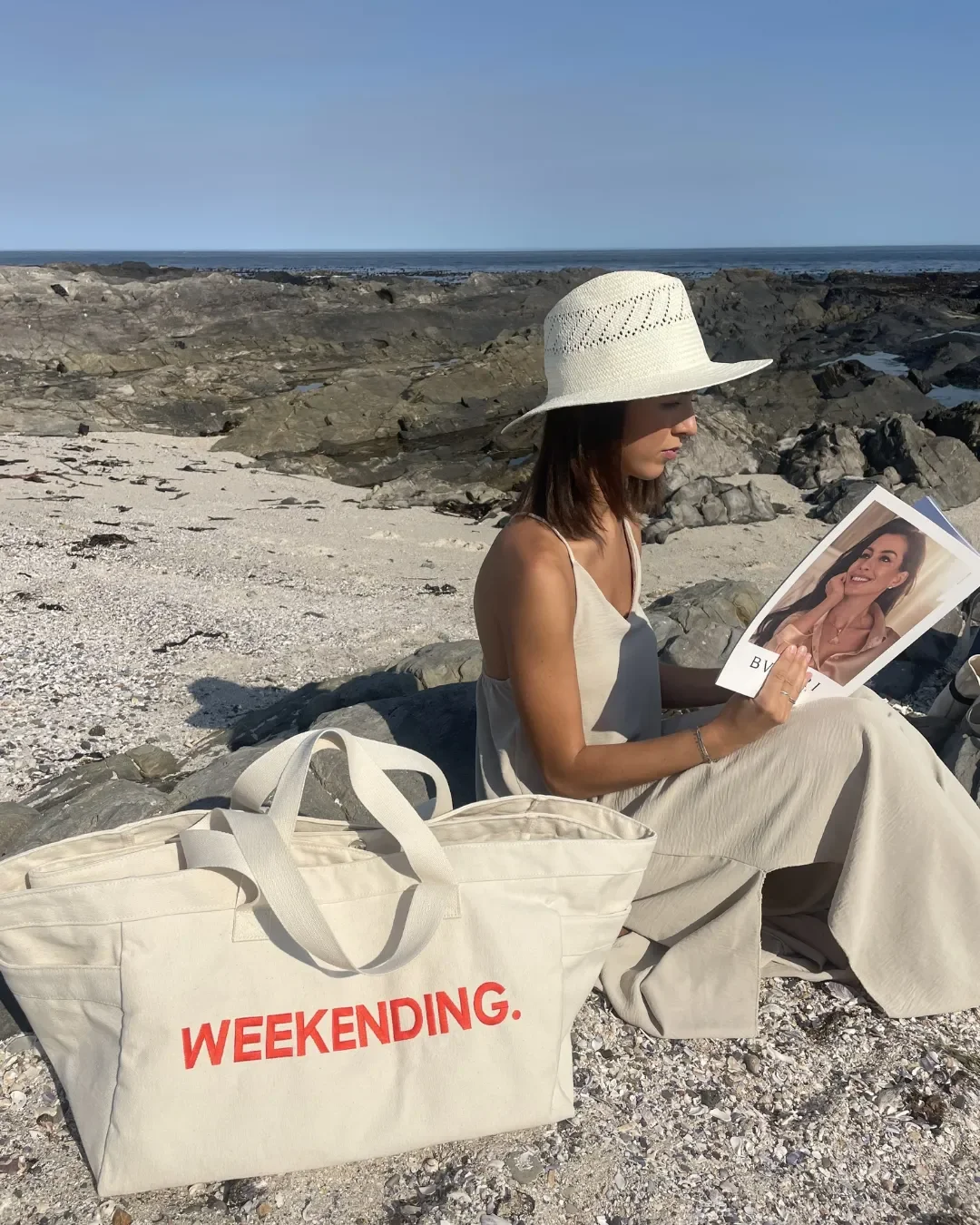 A woman sitting on a rocky beach reading a magazine next to a large cream 100% cotton canvas Weekending Bag with red printed branding, Handmade by Soli in Cape Town for merchandise or corporate gifts.