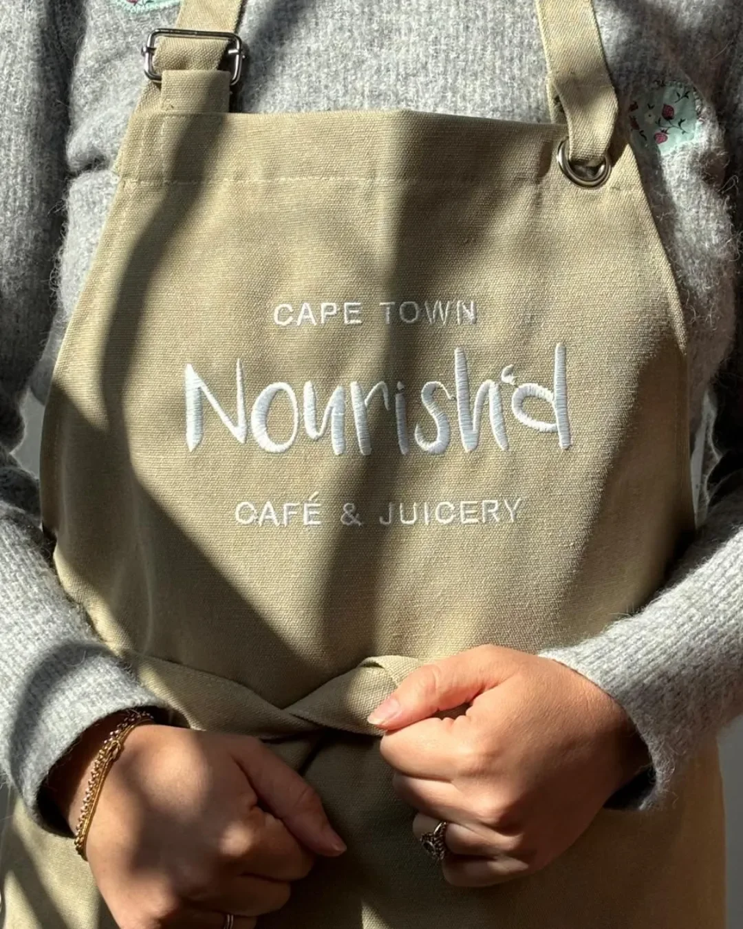 Close-up of custom white embroidered logo details on a beige canvas apron chest, made for Nourish'd Café & Juicery in Cape Town by Soli.