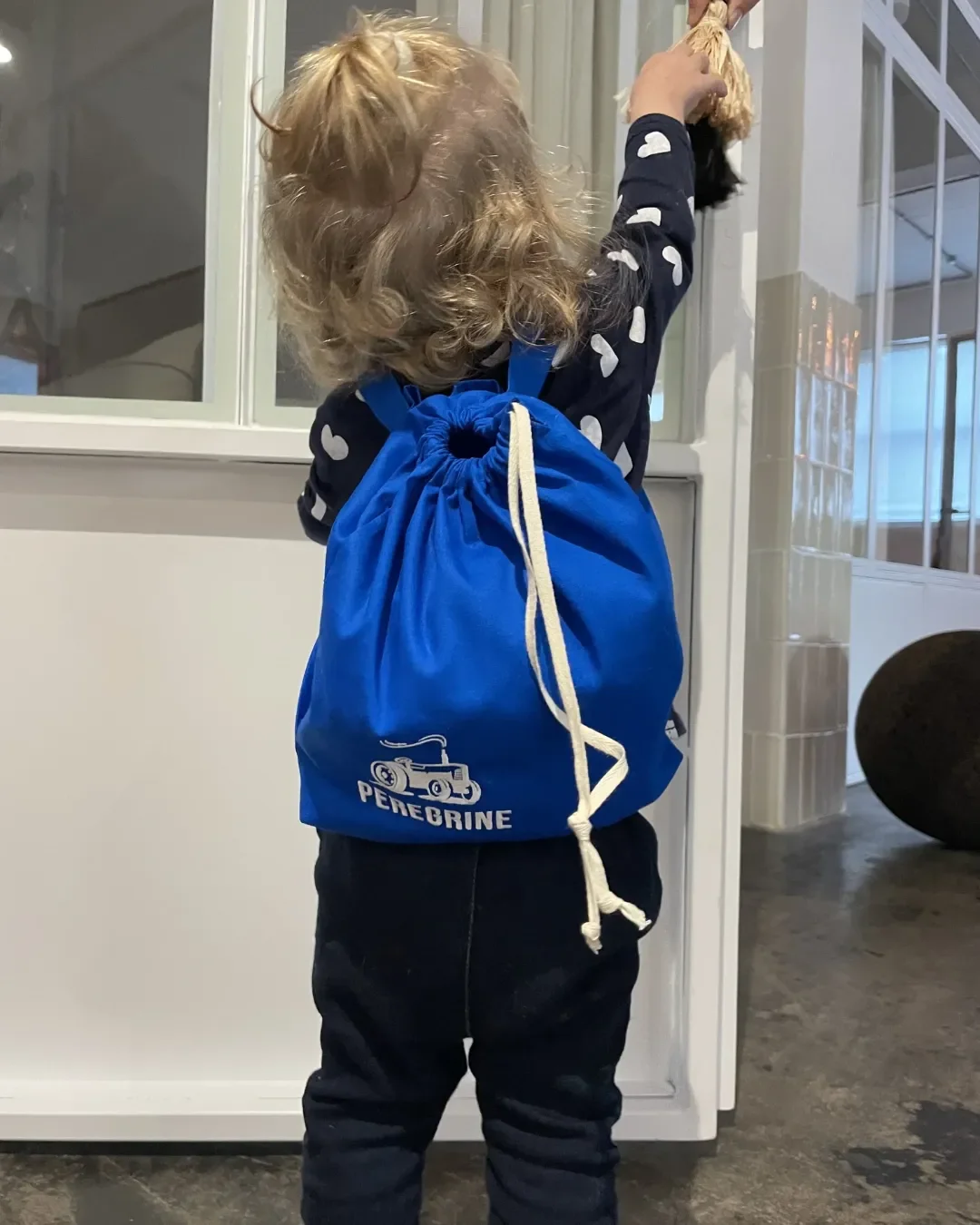 Images showing a toddler wearing the Soli Mini Backpack blue, featuring a custom printed tractor logo and adjustable drawstring straps. Made in Cape Town for corporate gifts and merchandise