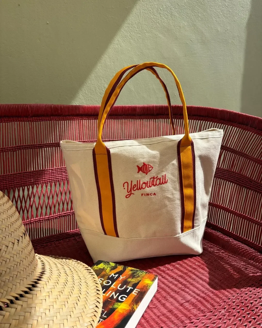Cream cotton canvas Oxford bag with custom embroidery and striped handles placed on a red wicker chair next to a straw hat. Handmade by Soli in Cape Town for sustainable merchandise or corporate gifts.
