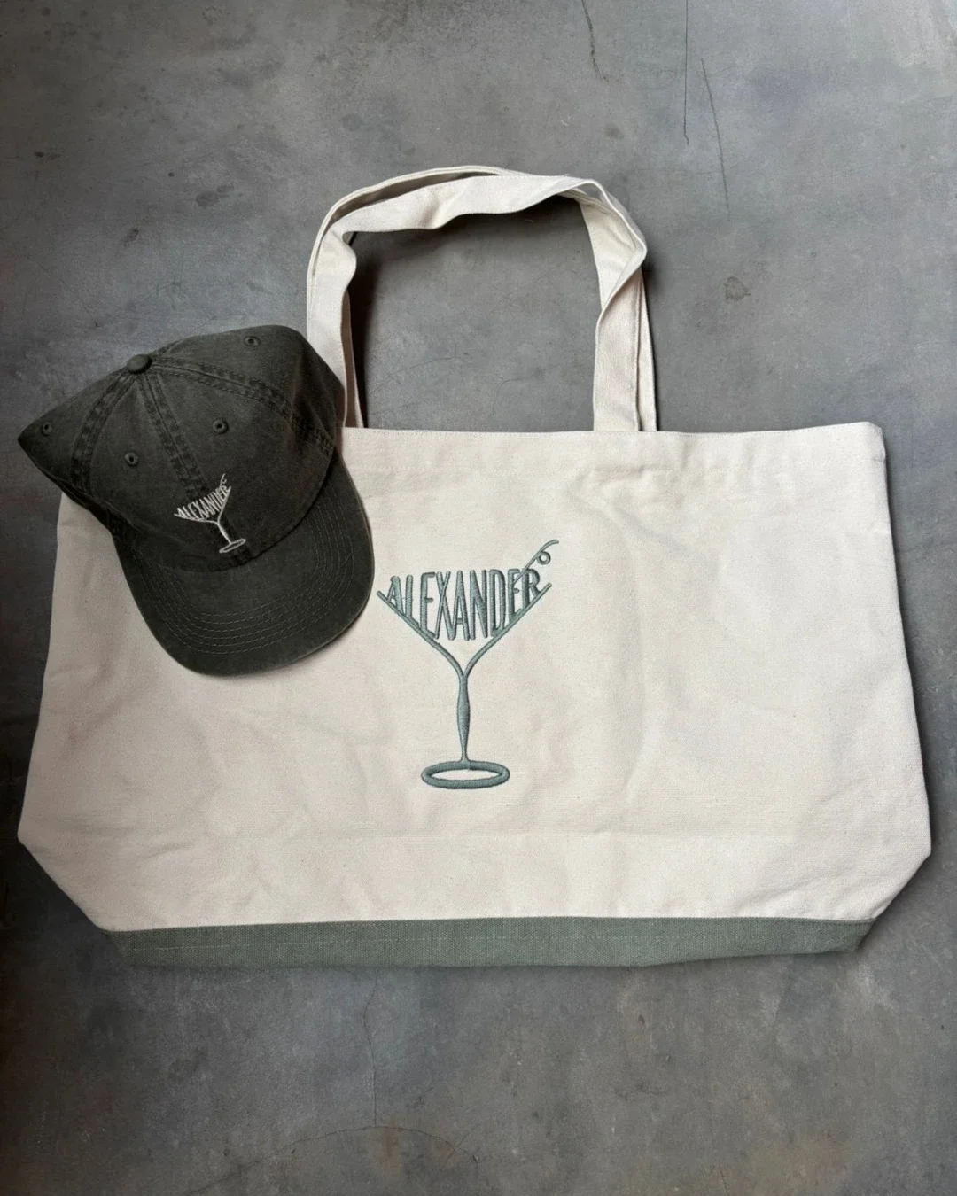 Cream cotton canvas Pezula bag and matching olive green cap featuring custom "ALEXANDER" martini glass embroidery, displayed on a textured grey concrete background, handmade by Soli in Cape Town for sustainable merchandise or corporate gifts.