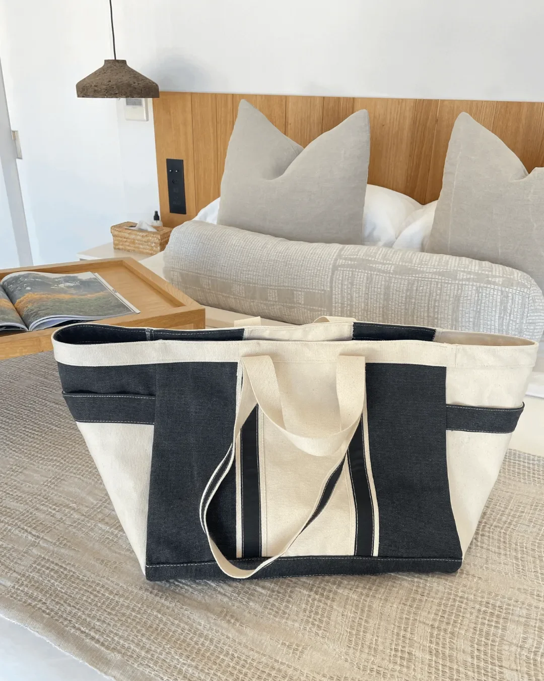 A large black and cream Midnight canvas tote bag resting on a bed in a modern hotel room, handmade by Soli in Cape Town.