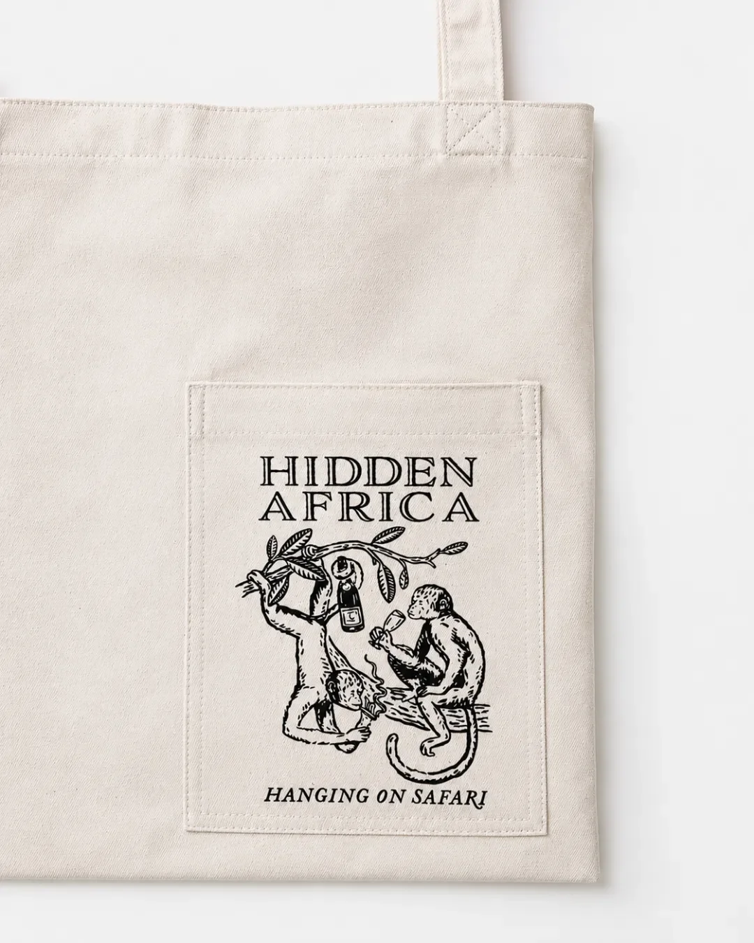 Close-up of a cream canvas tote bag's stitched front pocket, showing a black line-art graphic of monkeys drinking wine with the text "HIDDEN AFRICA HANGING ON SAFARI".