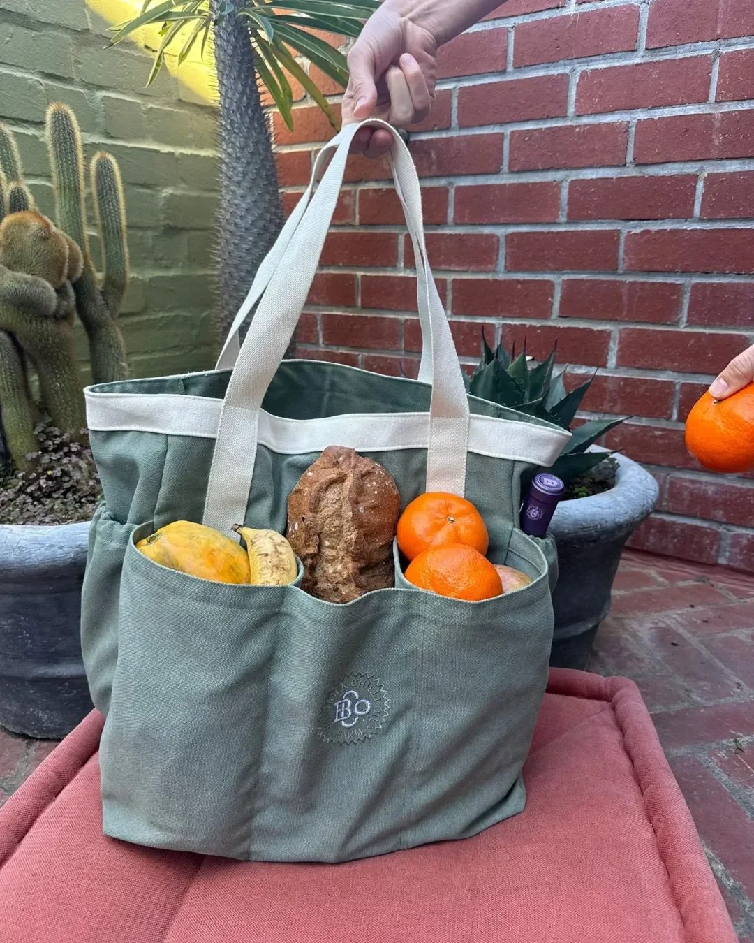 An olive green 100% canvas Market Bag with multiple exterior pockets filled with fresh bread and fruit, featuring white contrast handles and 'BO' embroidery, Handmade by Soli in Cape Town for merchandise or corporate gifts.