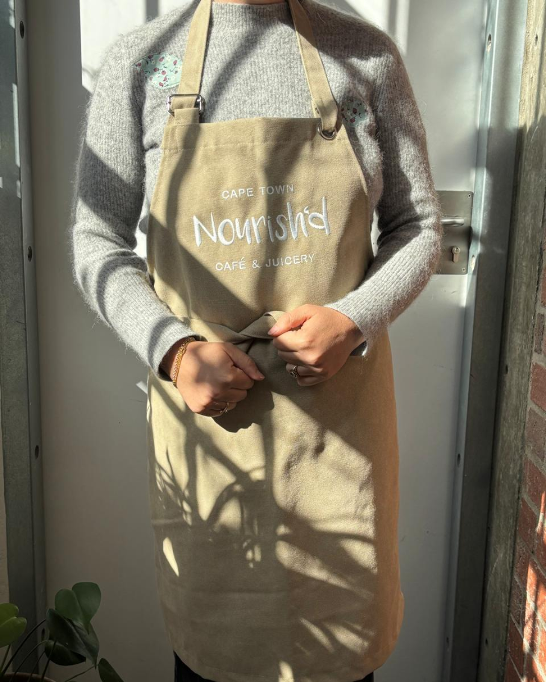 Front view of a person wearing a beige canvas full-body bib apron with custom white embroidery reading "CAPE TOWN Nourish'd CAFÉ & JUICERY", crafted by Soli.