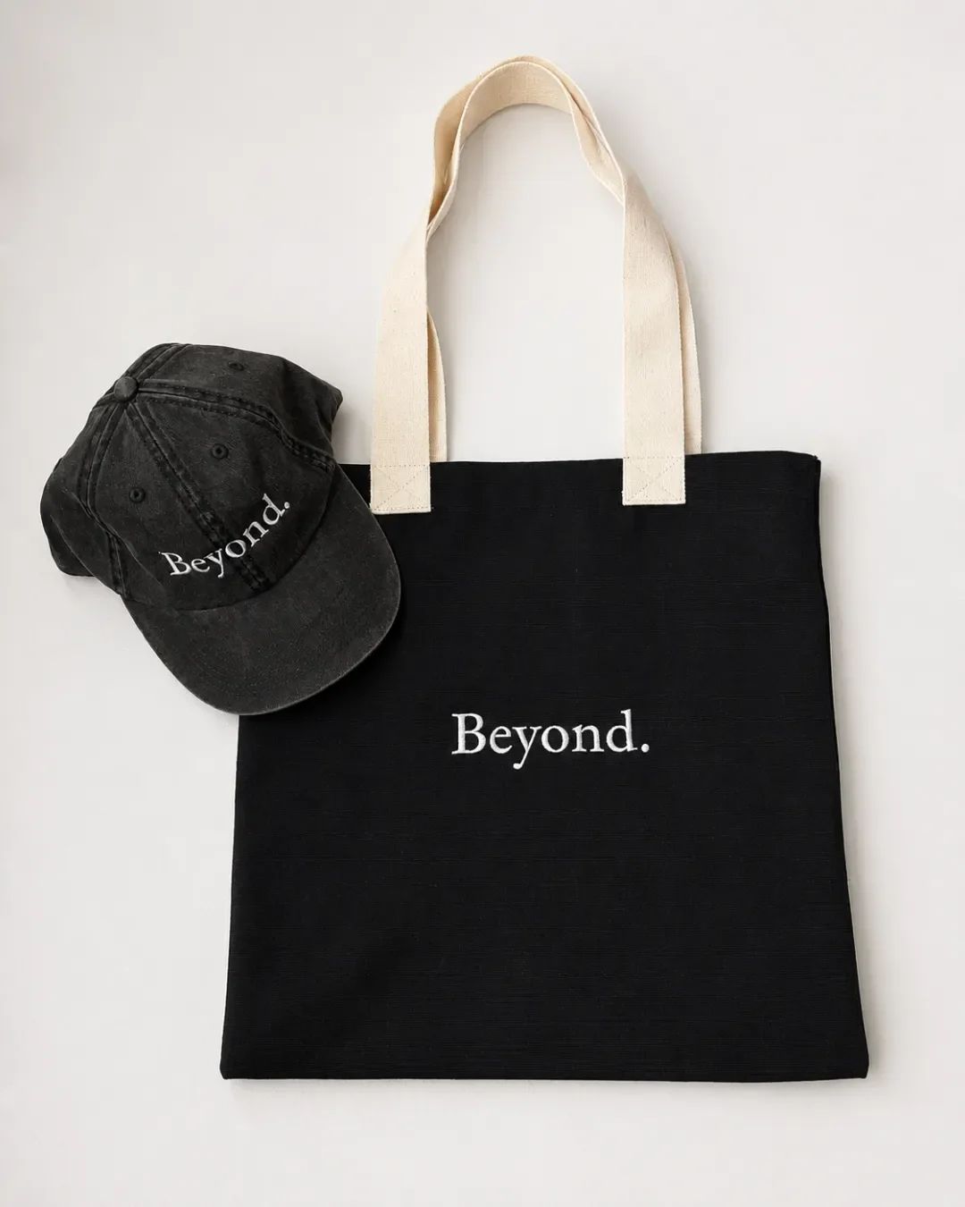Top-down overhead view of a custom black 2-tone canvas tote bag with cream straps and an embroidered logo next to a matching black cap, handmade by Soli in Cape Town for branded merchandise or corporate gifts.