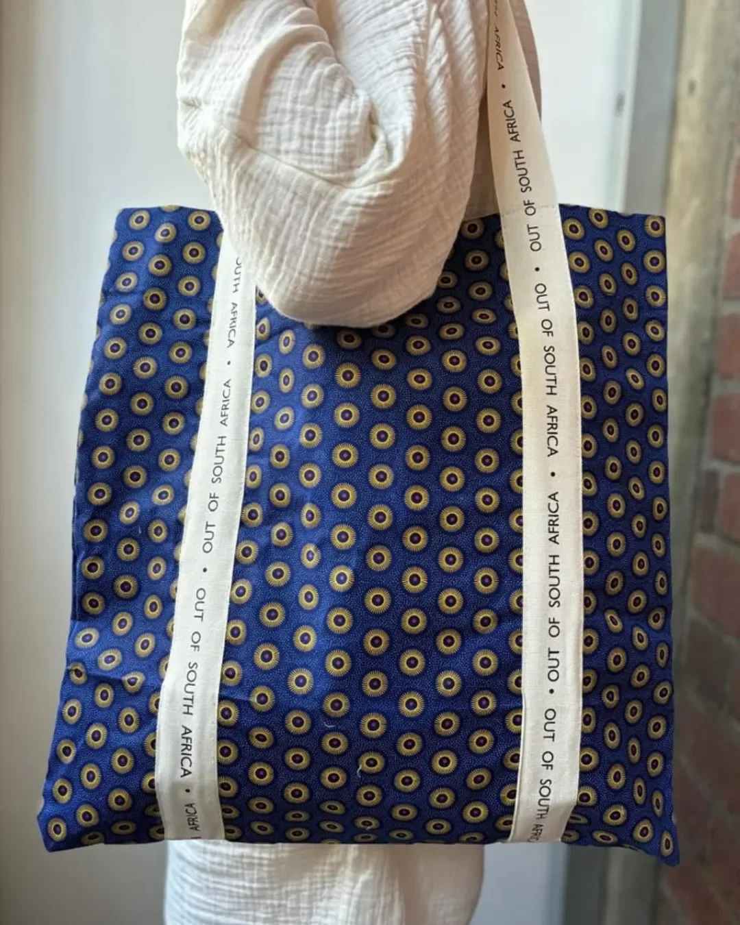 A person carrying a vibrant blue 100% shweshwe tote bag with a yellow and brown bullseye pattern, featuring long custom white cotton ribbons printed with 'OUT OF SOUTH AFRICA', handmade by local artisans for Soli in Cape Town.