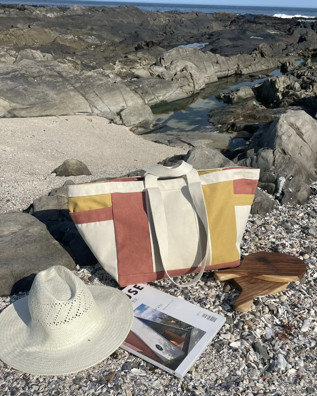 A multi-colored earth-toned canvas bag on a shell-covered beach with a sun hat and magazine, handmade by Soli in Cape Town.