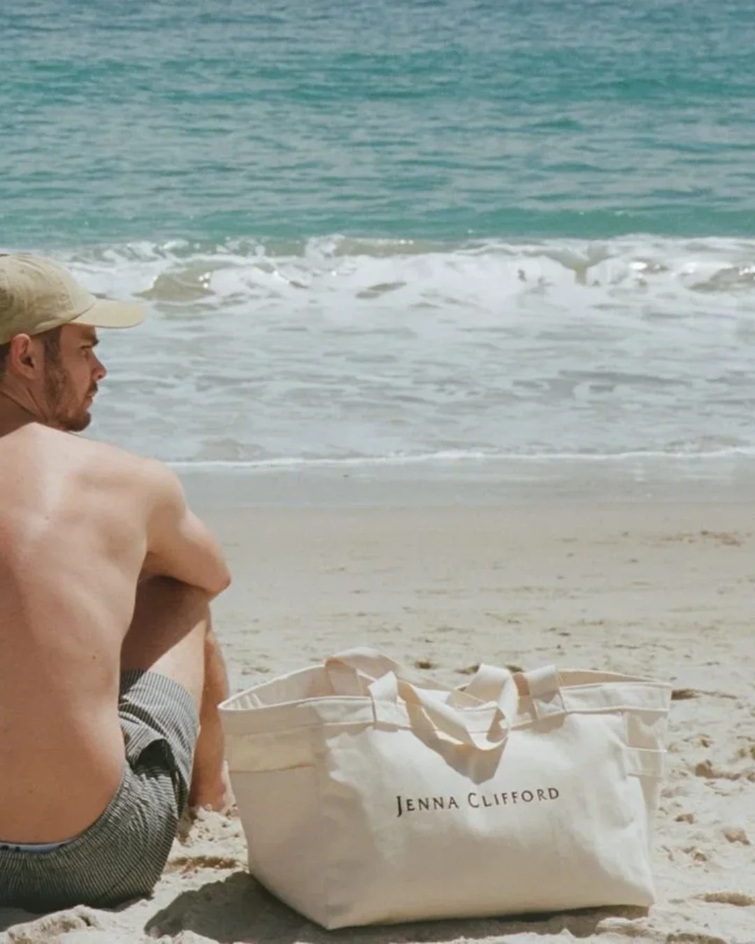 A large cream-colored 100% cotton canvas Weekending Bag with 'JENNA CLIFFORD' branding sitting on a sandy beach, Handmade by Soli in Cape Town for merchandise or corporate gifts.