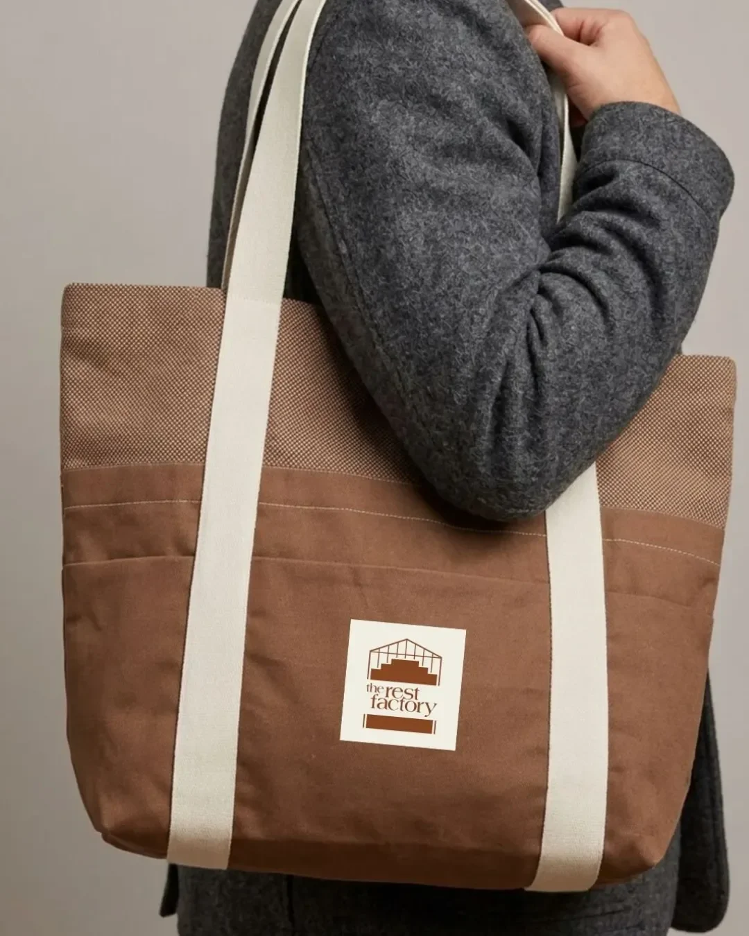 A brown canvas tote bag with contrasting cream shoulder straps, featuring a printed square logo for The Rest Factory, carried by a person wearing a textured grey wool coat for premium brand merchandise inspiration.