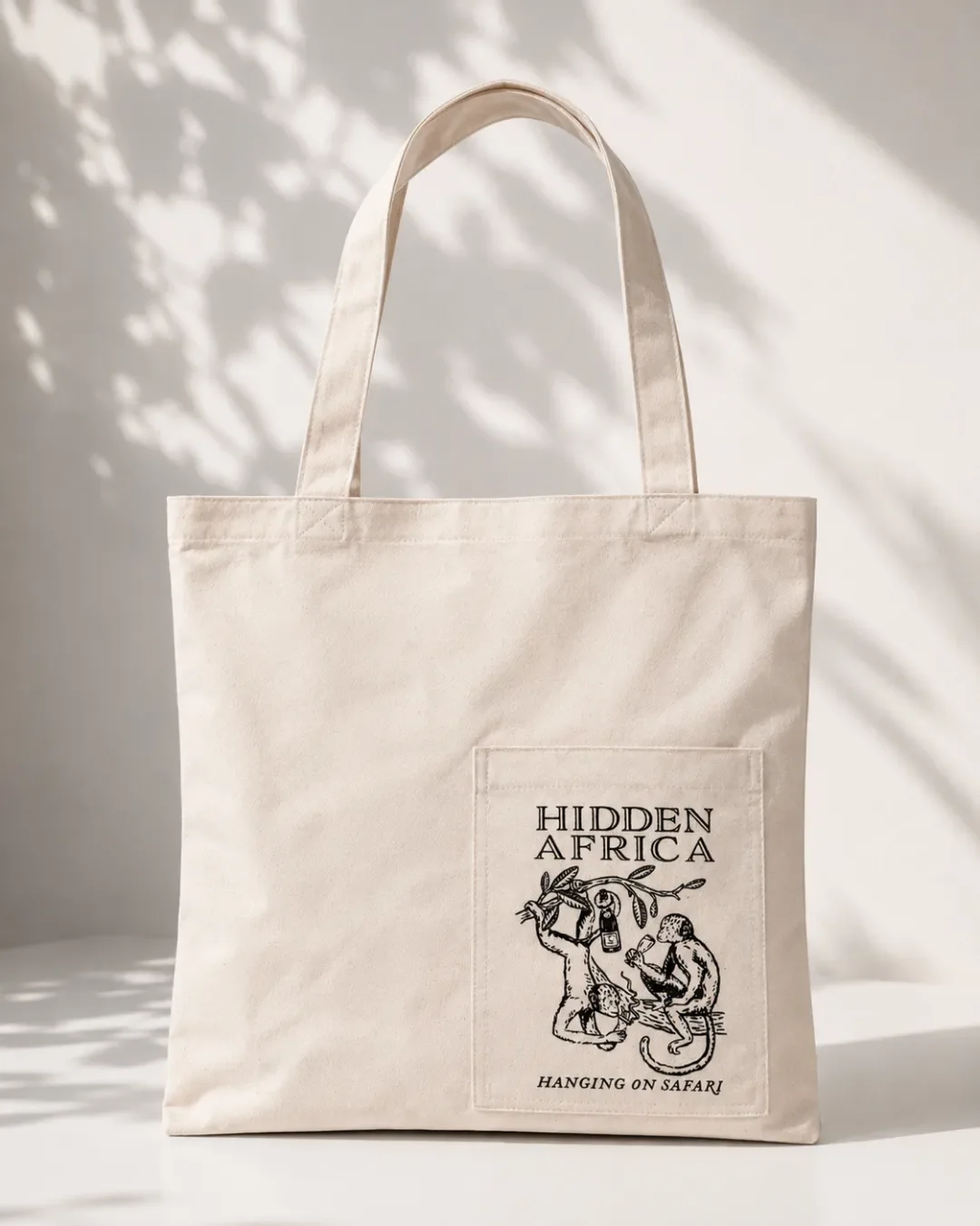 A cream canvas tote bag with an external front pocket featuring a black graphic illustration of monkeys and the text "HIDDEN AFRICA HANGING ON SAFARI", set against a neutral background with soft leaf shadows.