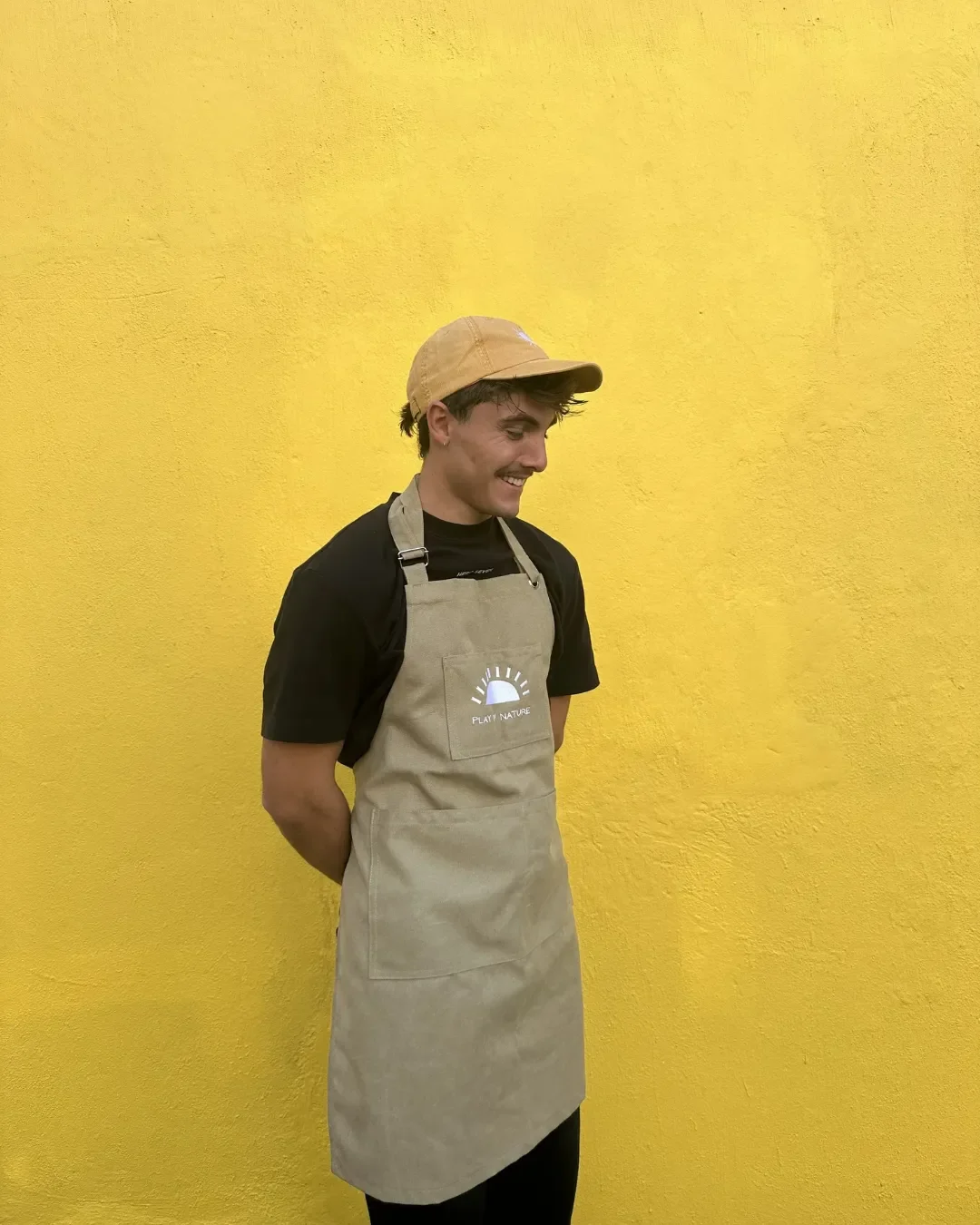 A man wearing a custom beige canvas full-body apron with a white embroidered logo and a matching cap, standing against a bright yellow wall. Made by Soli in Cape Town as a hospitality merchandise apron for staff.