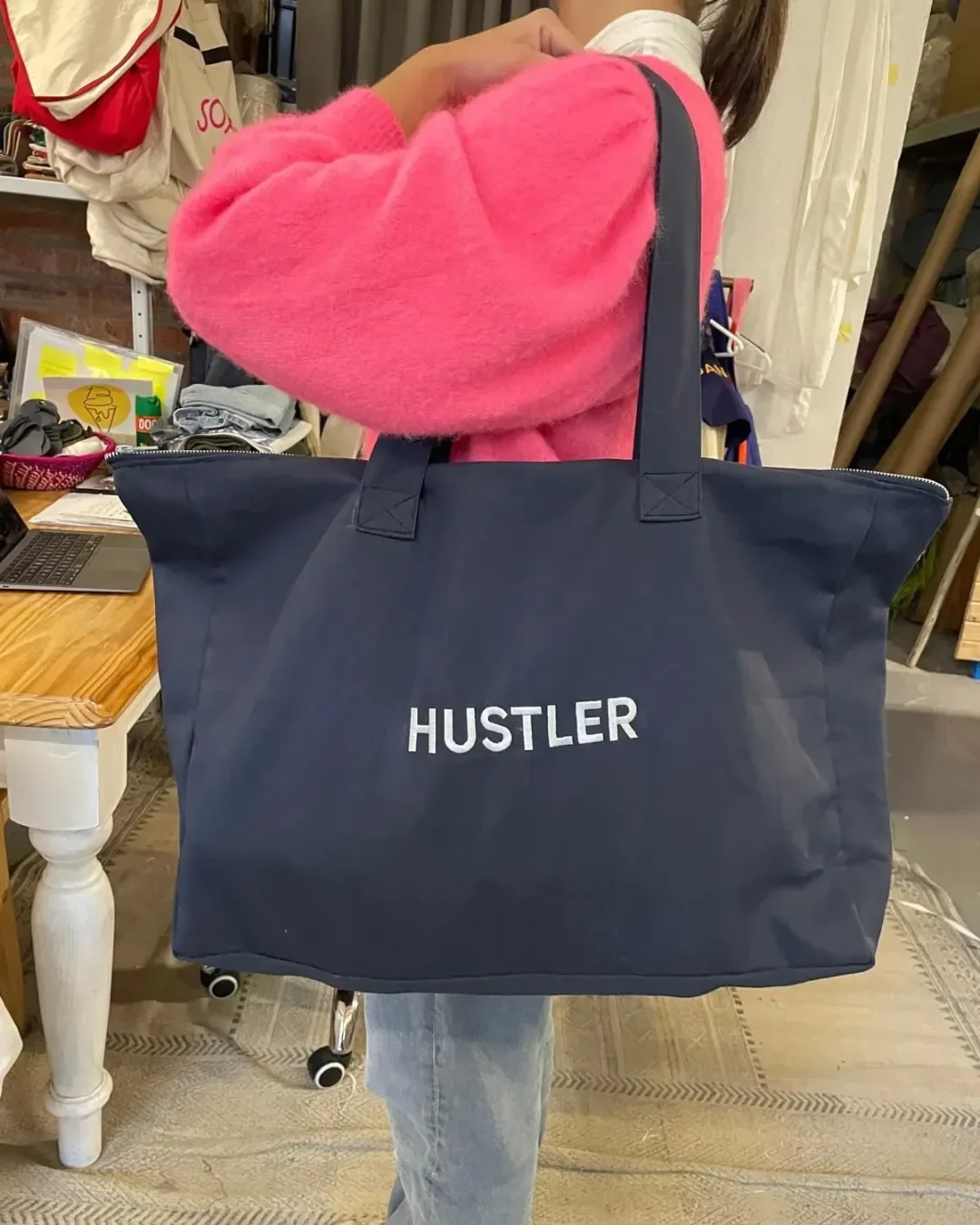 A large navy blue canvas tote bag with a top zip closure, branded with white "HUSTLER" text, held by a person wearing light wash jeans and a pink sweater in a workspace for custom corporate merchandise.
