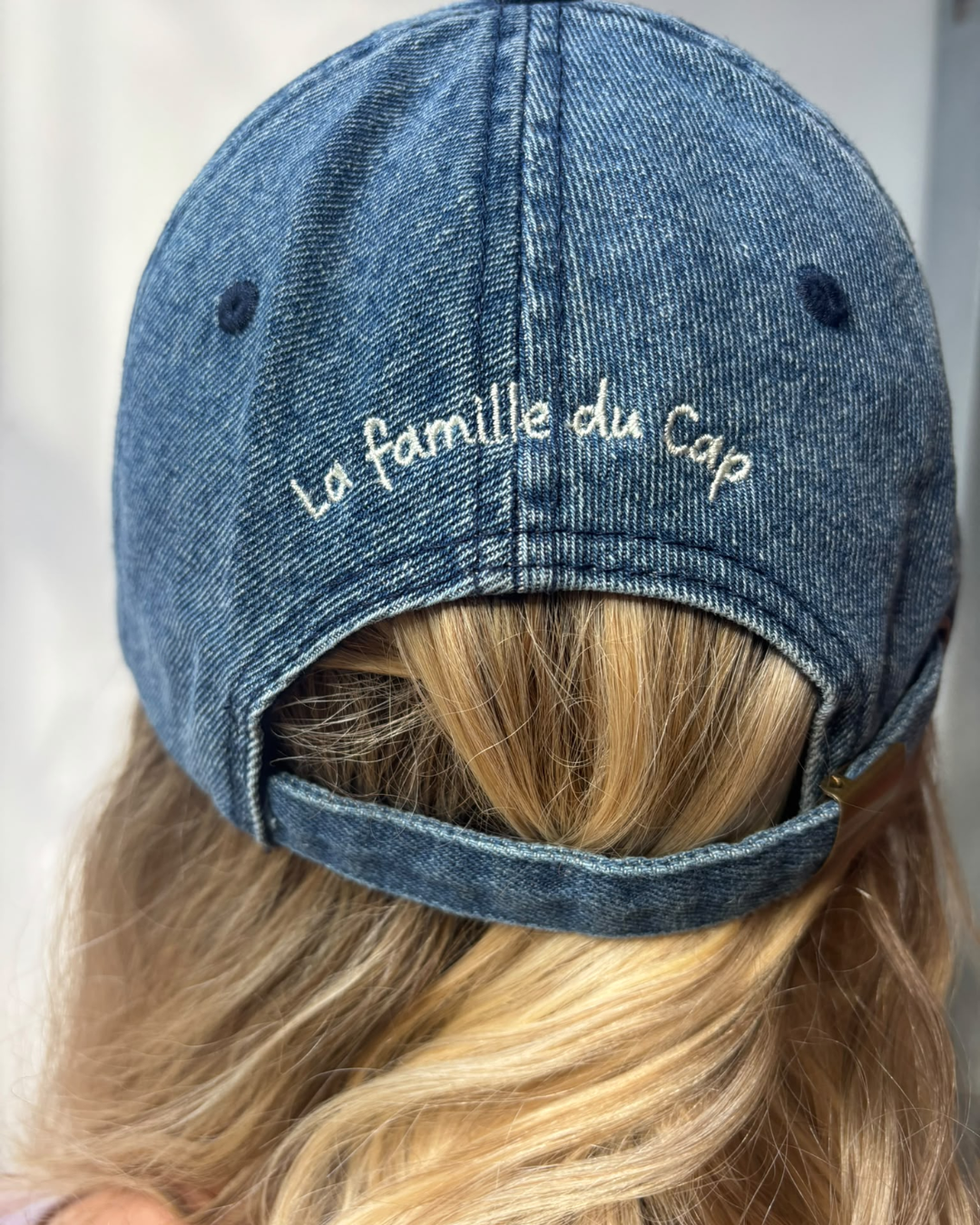 Close-up of the back of a washed denim baseball cap worn by someone with blonde hair, showcasing white hand-embroidered cursive text reading "La famille du Cap" above an adjustable strap.