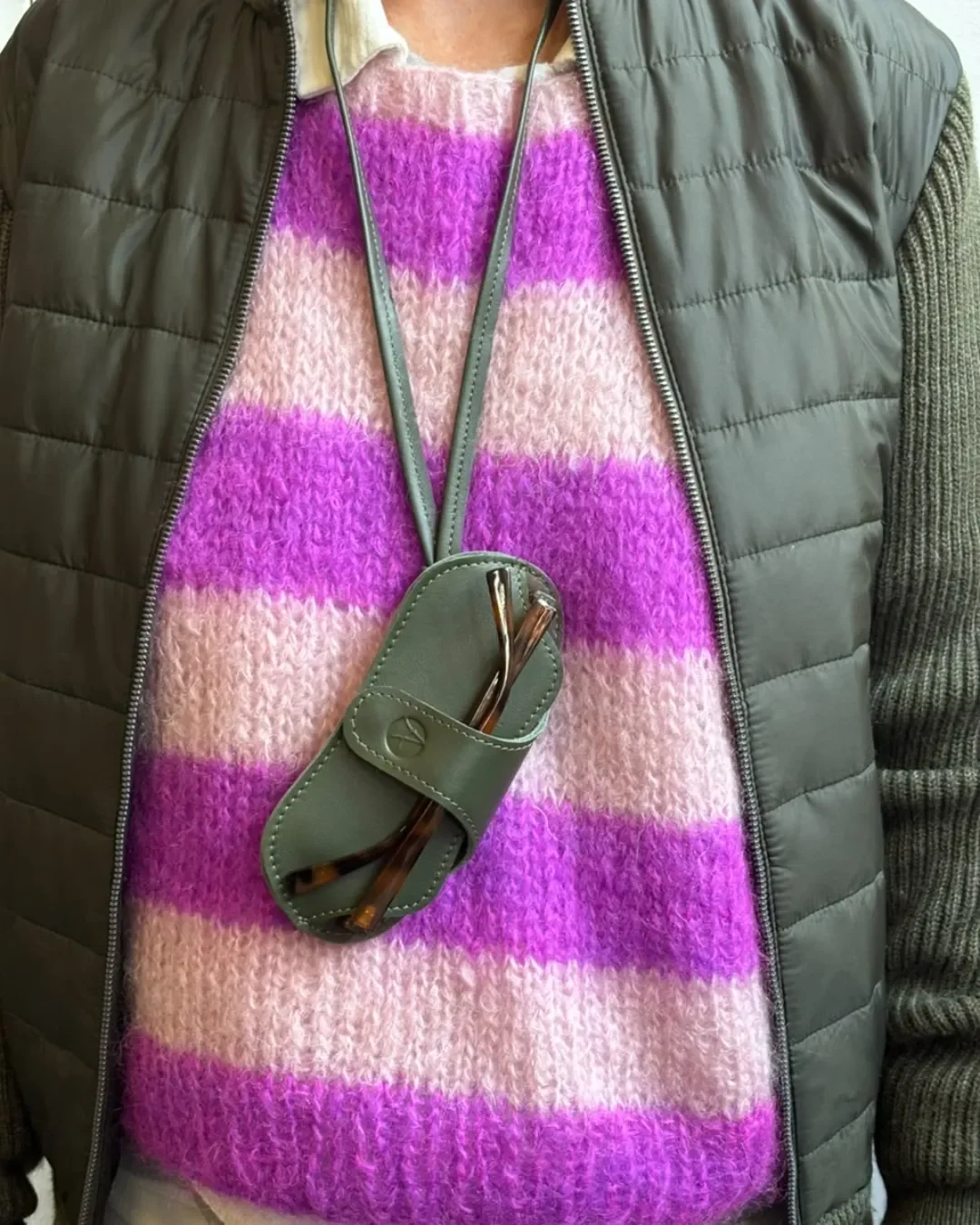Person wearing an olive green leather sunglasses pouch lanyard over a sweater, holding tortoise shell glasses, custom branded by Soli.