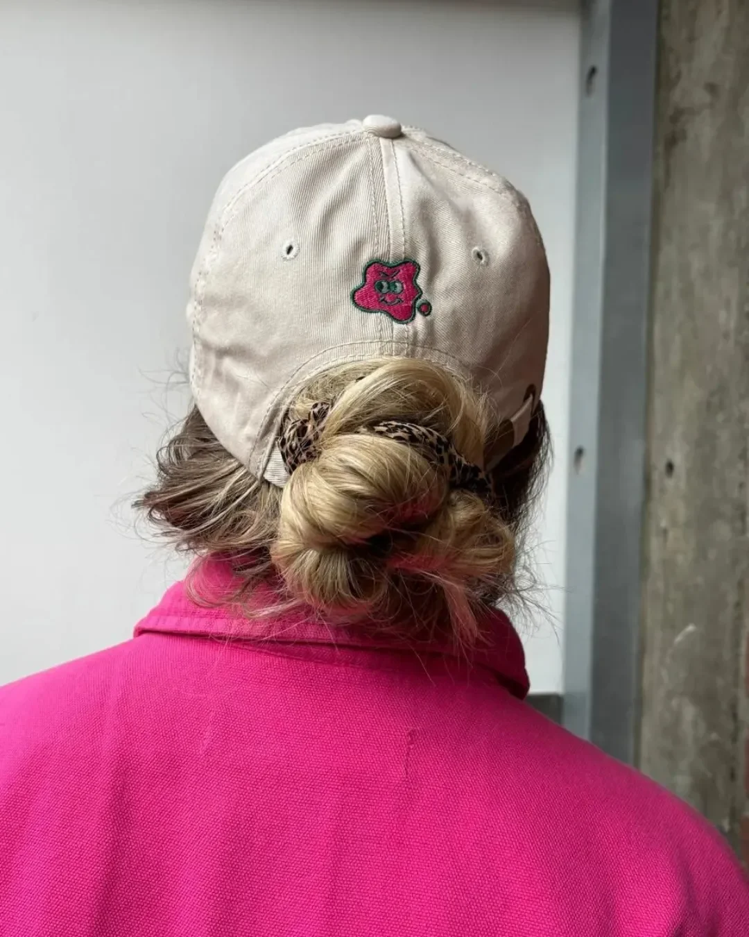 Rear view of a custom cream-colored dad cap featuring a small pink and green cartoon character embroidered on the back, worn by a person with their hair styled in a bun with a leopard print scrunchie, paired with a bright pink jacket.