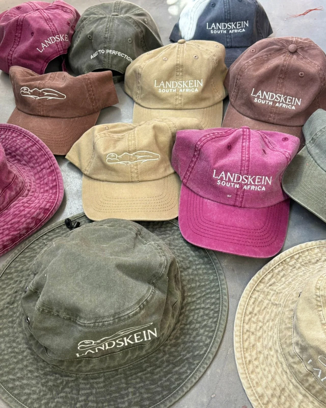 An assortment of washed canvas custom caps, bucket hats, and wide-brim safari hats in earthy tones like olive green, khaki, brown, and pink, embroidered with the Landskein South Africa branding for premium promotional merchandise.