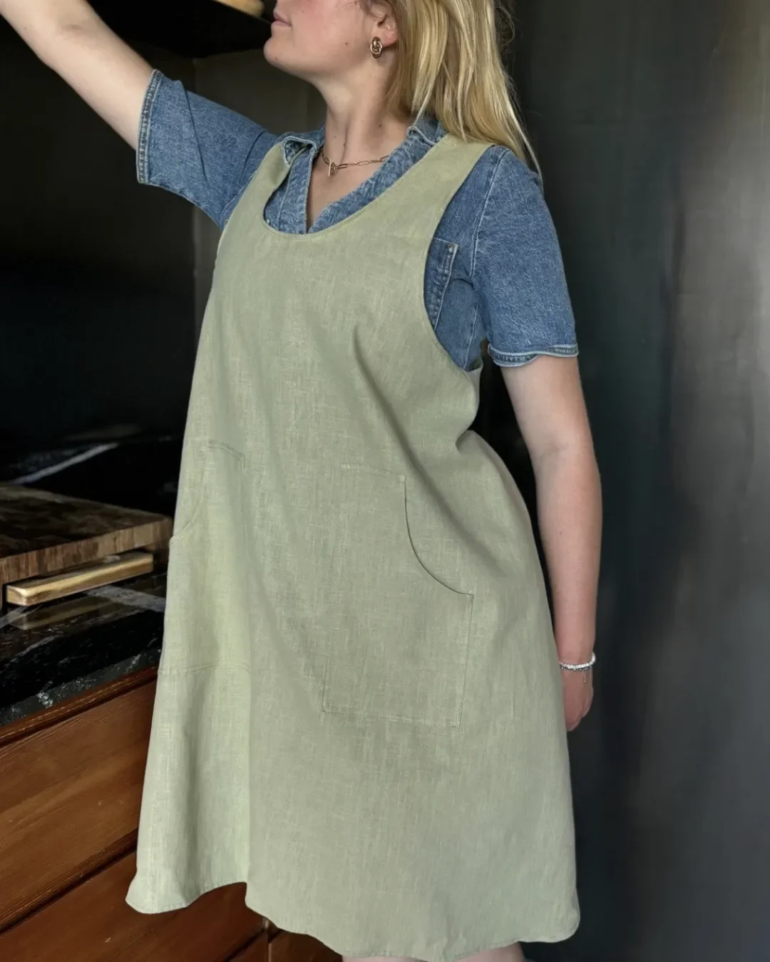 A woman wearing a sage green ramie farmer apron with large front pockets over a denim shirt, handmade by Soli in Cape Town for Hospitality staff merchandise.