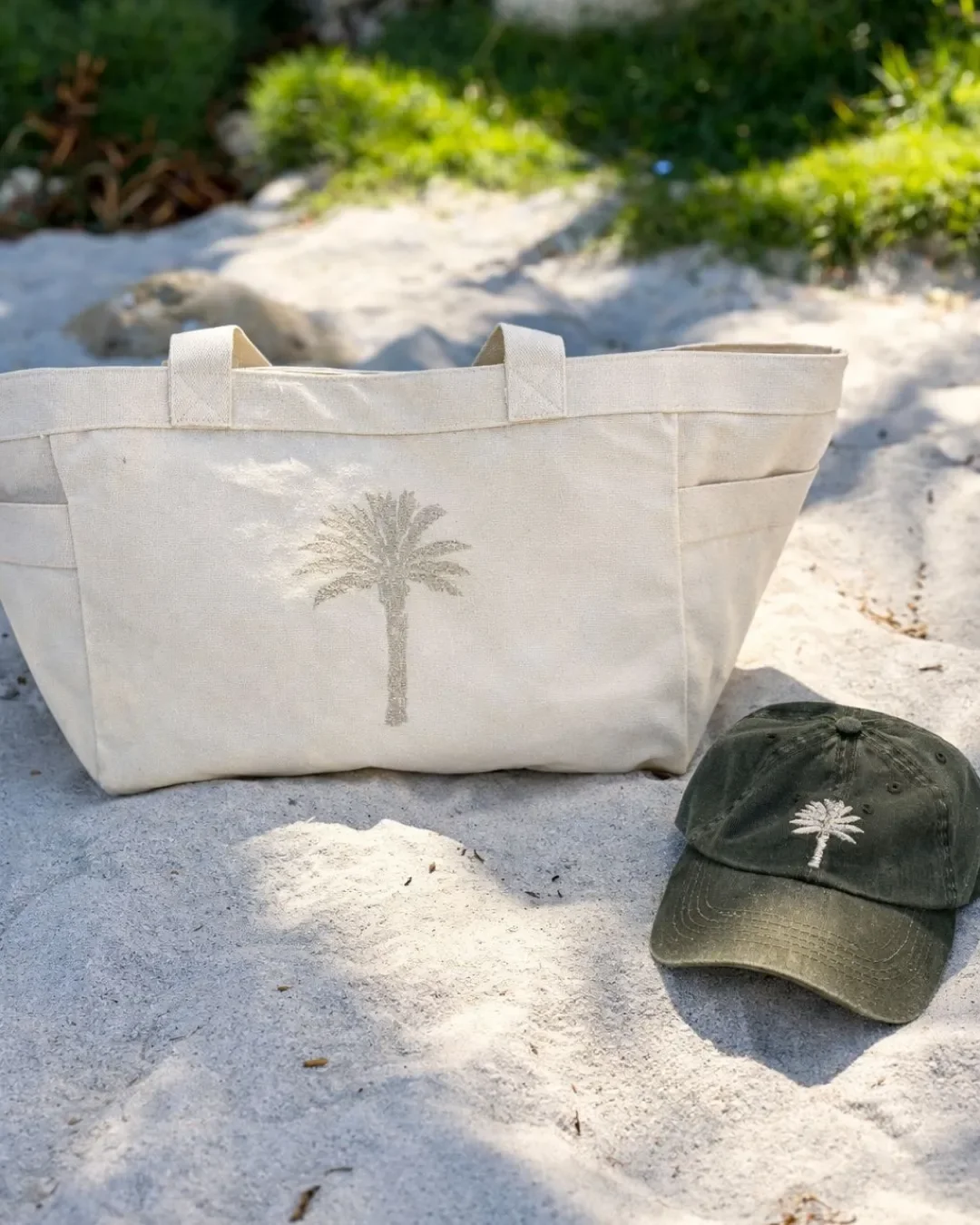 A large cream-colored 100% cotton canvas Weekending Bag with an embroidered palm tree logo and side pockets sitting on a sandy beach next to an olive green cap, Handmade by Soli in Cape Town for merchandise or corporate gifts.