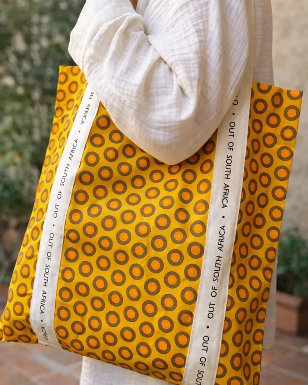 A person carrying a vibrant yellow 100% shweshwe tote bag with a circular bullseye pattern, featuring long custom white cotton ribbons printed with 'OUT OF SOUTH AFRICA', handmade by local artisans for Soli in Cape Town.