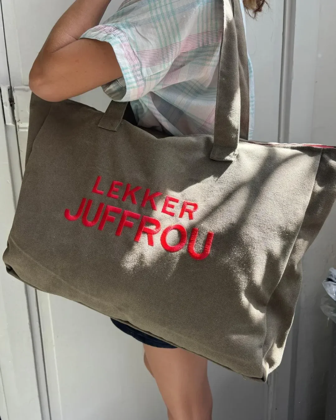 Close up of a person carrying a large olive green canvas tote bag with red Lekker Juffrou embroidery, Handmade by Soli in Cape Town for sustainable merchandise or corporate gifts.