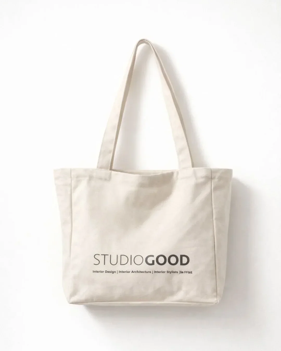 A cream canvas corporate tote bag featuring a minimalist printed logo for Studio Goodd, hanging against a clean white wall as an example of premium custom merchandise.