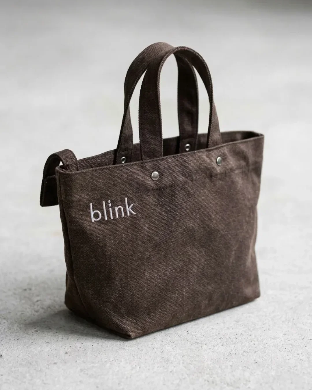 A textured brown heavy-duty fabric Japanese-style wrap bag featuring white "blink" embroidery and silver stud detailing on a grey concrete background, handmade by Soli in Cape Town for sustainable merchandise or corporate gifts.