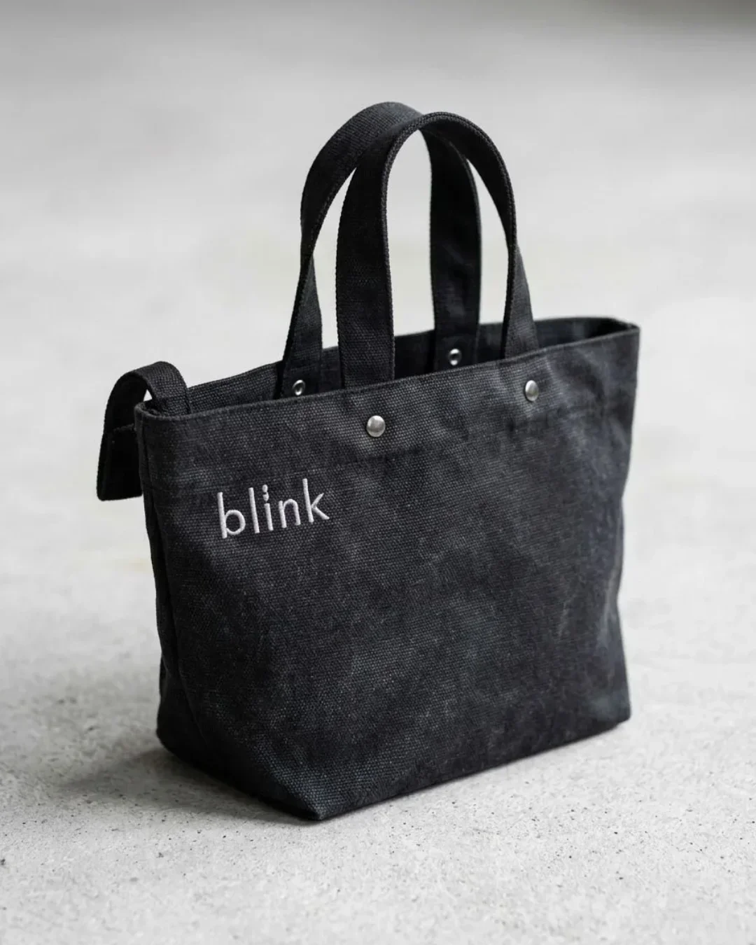 A charcoal grey Japanese-style wrap bag made from heavy-duty fabric featuring white "blink" embroidery and silver stud detailing on a textured grey background, handmade by Soli in Cape Town for sustainable merchandise or corporate gifts.