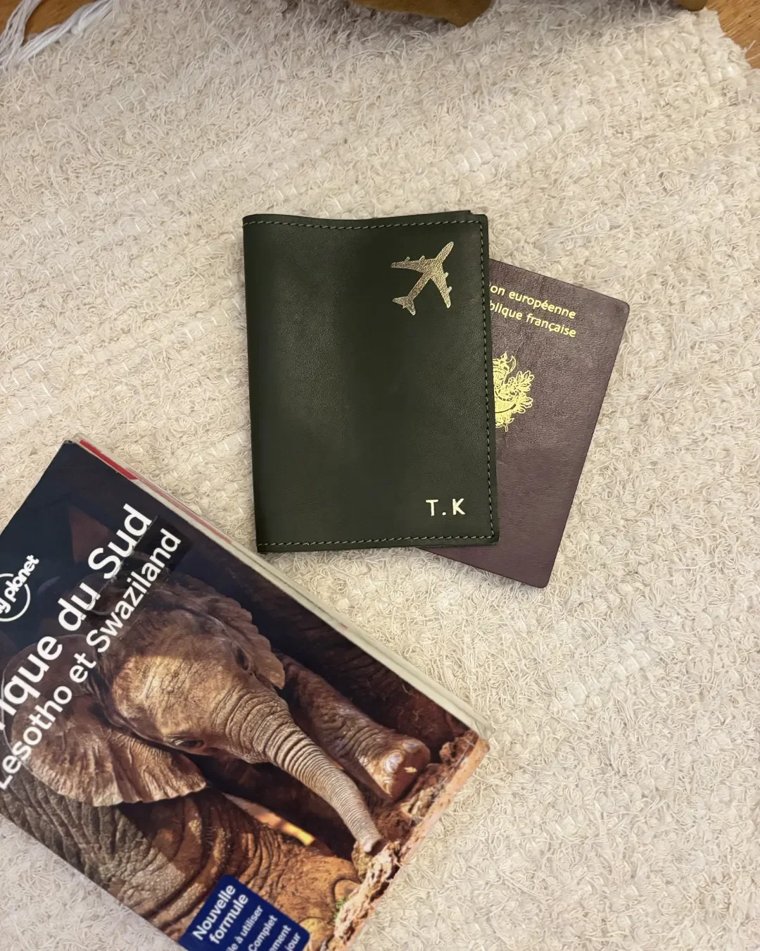 A custom olive green leather passport holder with gold-foiled airplane icon and "T.K" initials next to a French passport and travel book, Handmade by Soli in Cape Town for sustainable merchandise or corporate gifts.