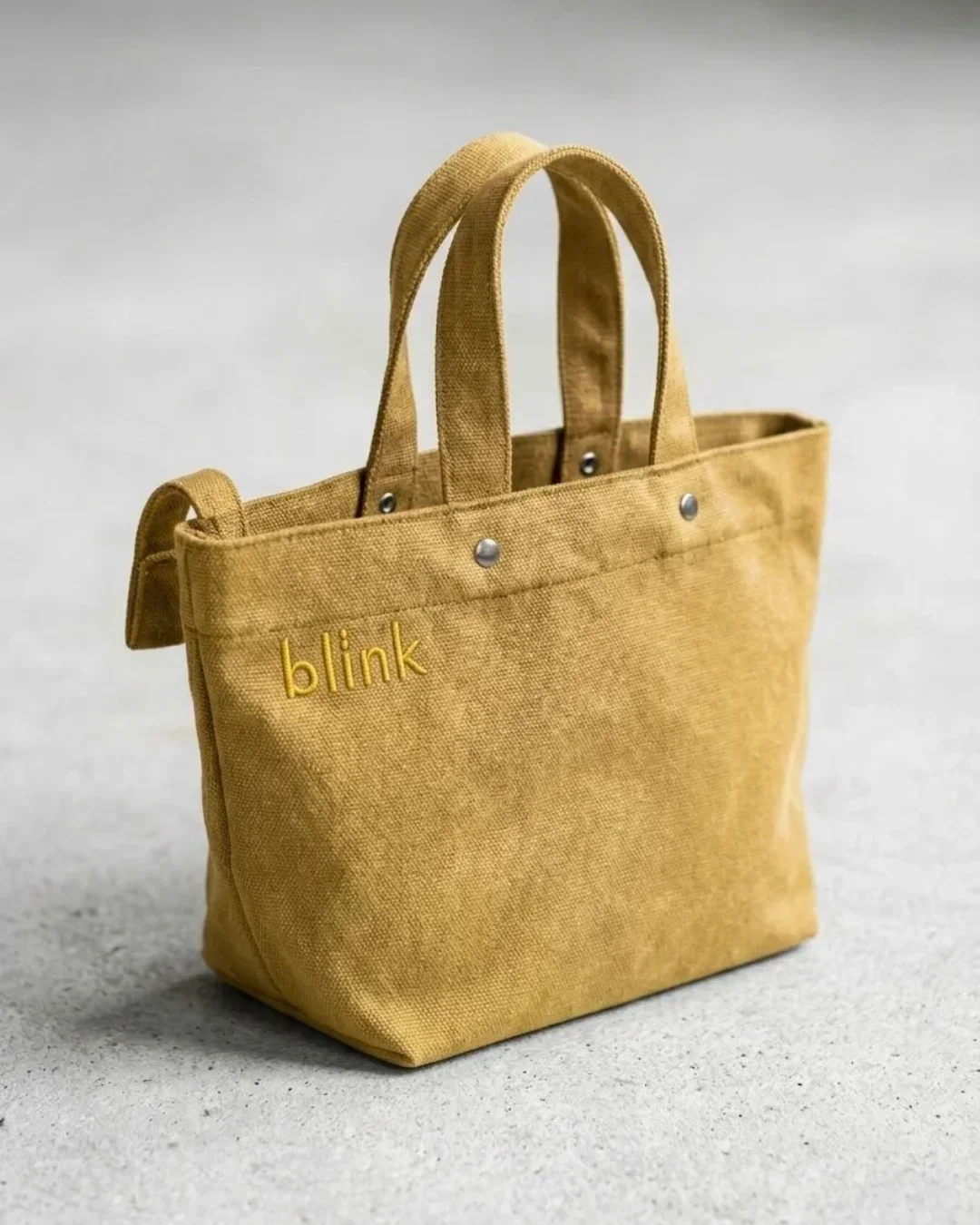 A mustard yellow Japanese-style wrap bag made from heavy-duty fabric featuring tonal "blink" embroidery and silver stud detailing on a textured grey background, handmade by Soli in Cape Town for sustainable merchandise or corporate gifts.