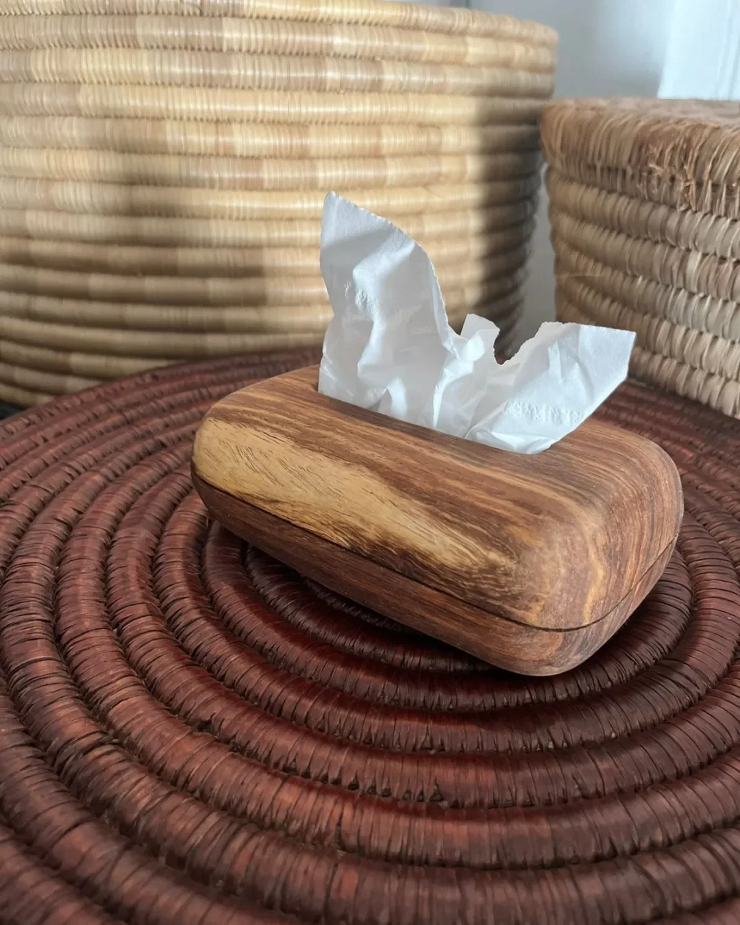 Handcrafted small Kiaat wood tissue box with rounded edges and natural grain, displayed on a woven African basket lid for luxury hotel room decor, by Soli made in Cape Town. 