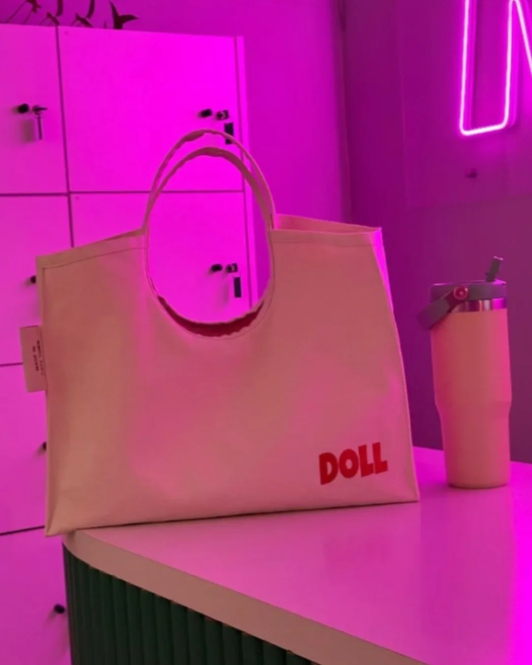 A cream heavy canvas Doll Bag featuring a rounded cotton handle and bold red "DOLL" embroidery, displayed on a white table in a room with vibrant pink neon lighting next to a reusable water bottle, handmade by Soli in Cape Town for sustainable mercha