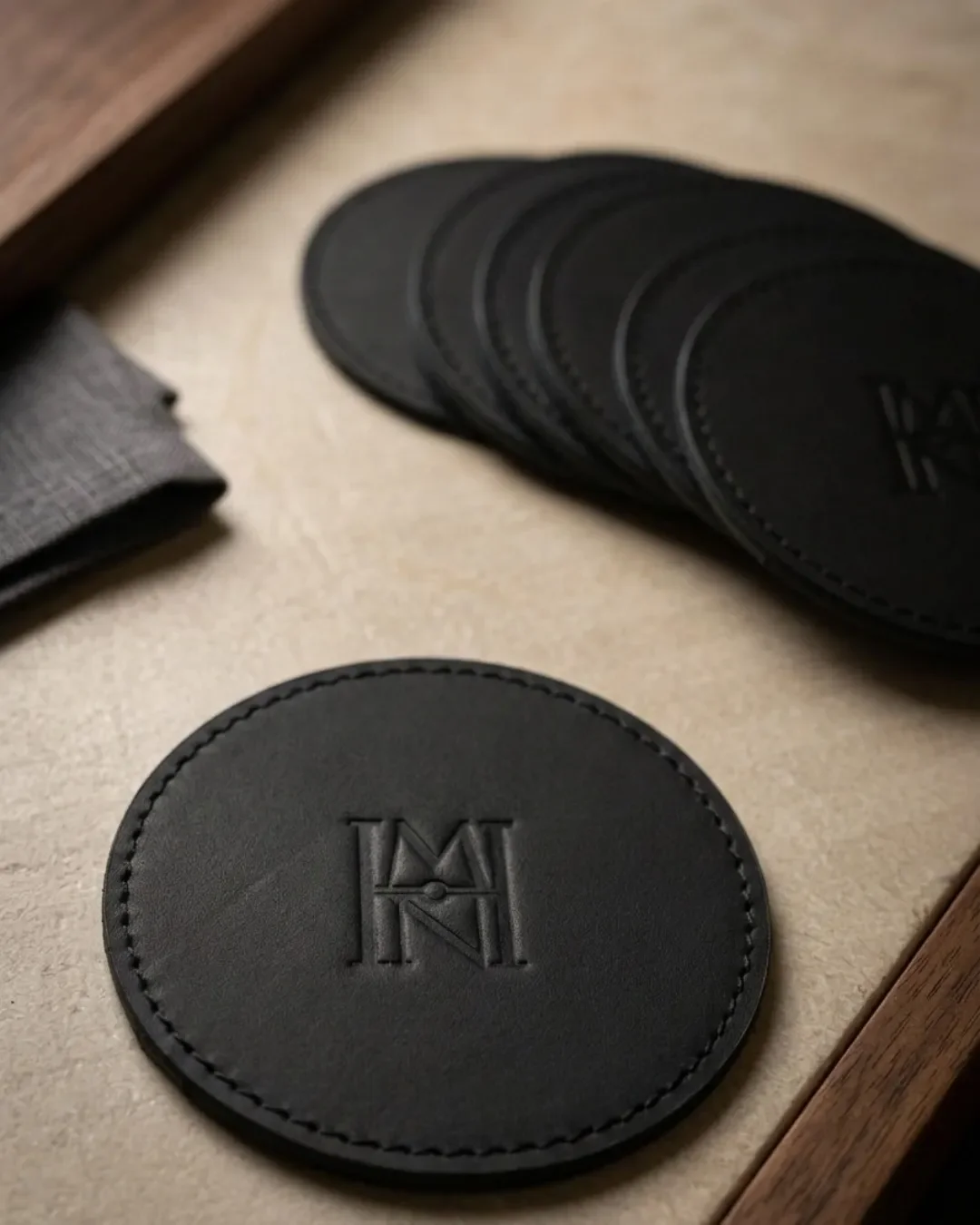 A detailed close-up of a handmade black leather coaster with a precisely debossed custom logo, surrounded by a stack of matching coasters and artisanal table settings. Coaster merchandise for hospitality made by Soli in Cape Town using sustainable ma