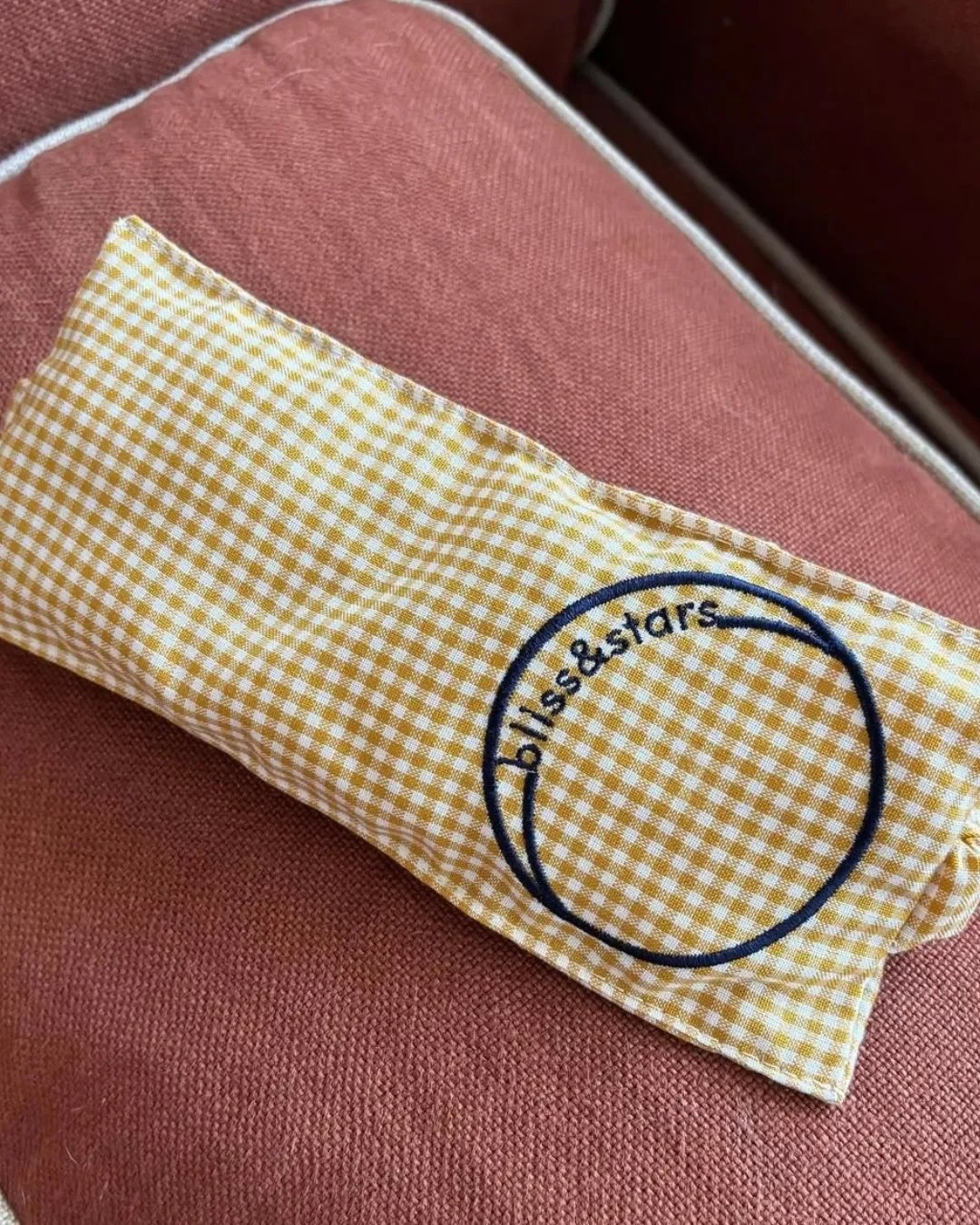 A yellow and white gingham check eye mask featuring a custom navy "bliss&stars" embroidered logo on a terracotta background, Handmade by Soli in Cape Town for sustainable merchandise or corporate gifts.