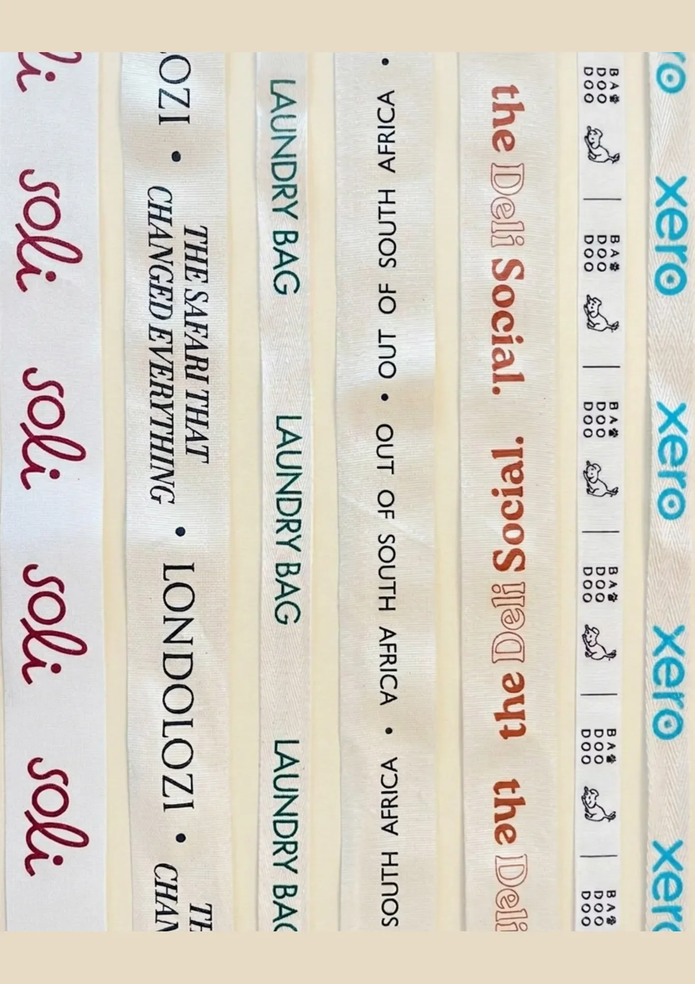 A vertical collection of custom printed cotton ribbon straps and branding tape displaying various corporate logos—including Soli, Londolozi, Xero, the Deli Social, and Ba Doo Doo—showcasing custom clothing ribbon labels and merchandise handles.