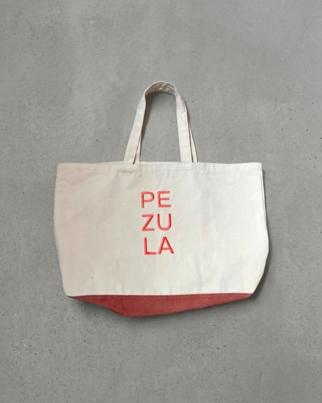 Cream 100% South African cotton canvas Pezula bag featuring a vibrant red contrasting base and matching red vertical "PEZULA" typography on a clean grey concrete floor, handmade by Soli in Cape Town for sustainable merchandise or corporate gifts.