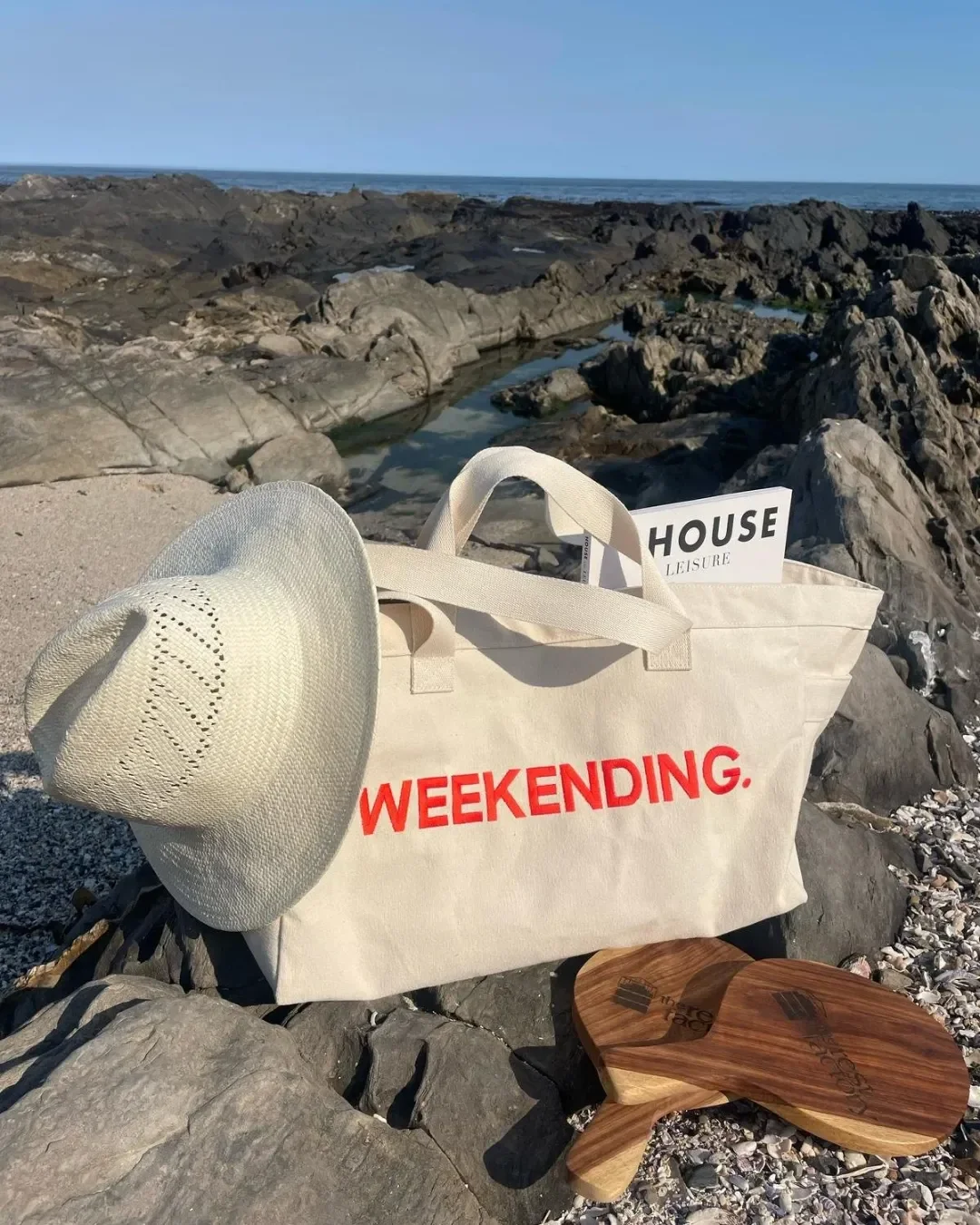 A large cream 100% cotton canvas Weekending Bag with red printed branding sitting on coastal rocks next to a straw hat and wooden boards, Handmade by Soli in Cape Town for merchandise or corporate gifts.