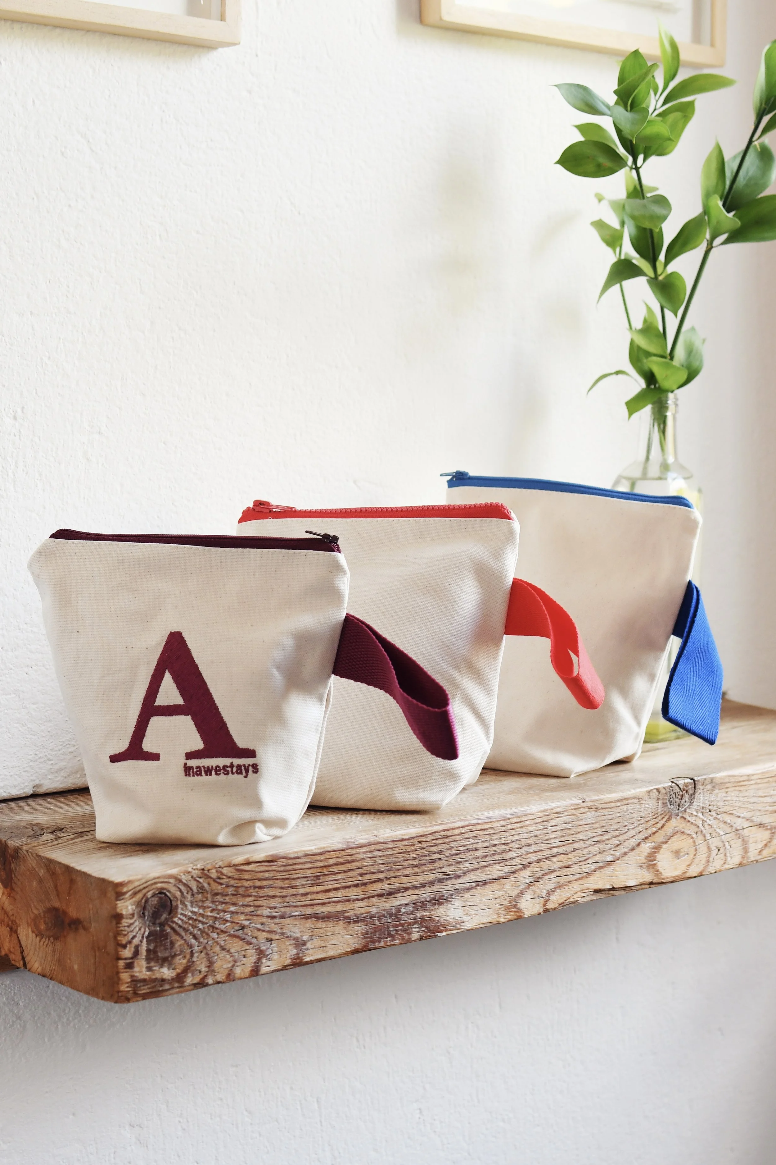 Three canvas pouches with zippers in different colors, displayed on a wooden shelf. The left pouch has a maroon letter 'A' and matching strap, the middle has a red strap, and the right one has a blue strap. A green leafy plant in a glass vase is part