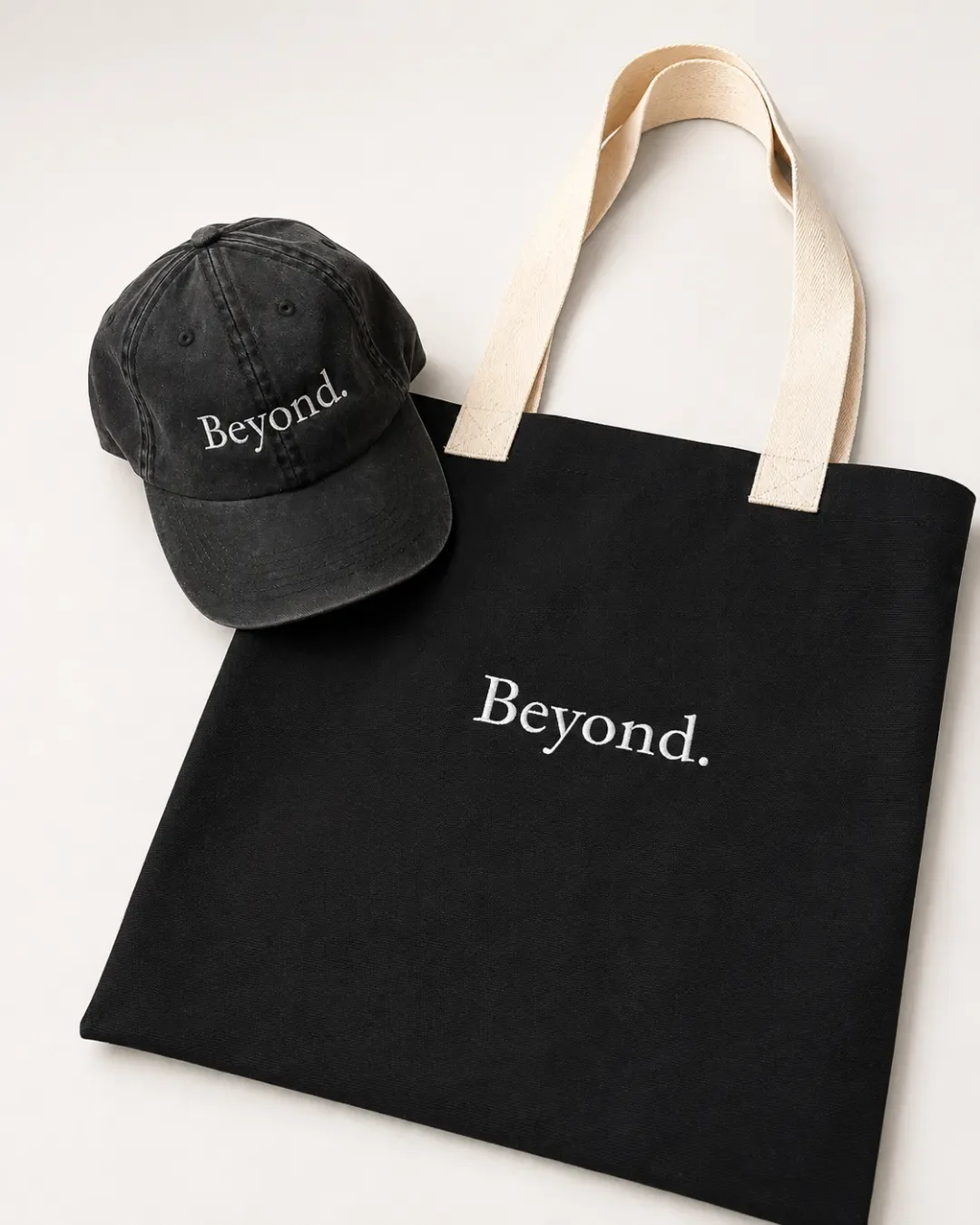 Angled close-up flat lay of a black canvas 2-tone tote bag with cream handles and white embroidered Beyond logo beside a washed black cap, handmade by Soli in Cape Town for branded merchandise or corporate gifts.